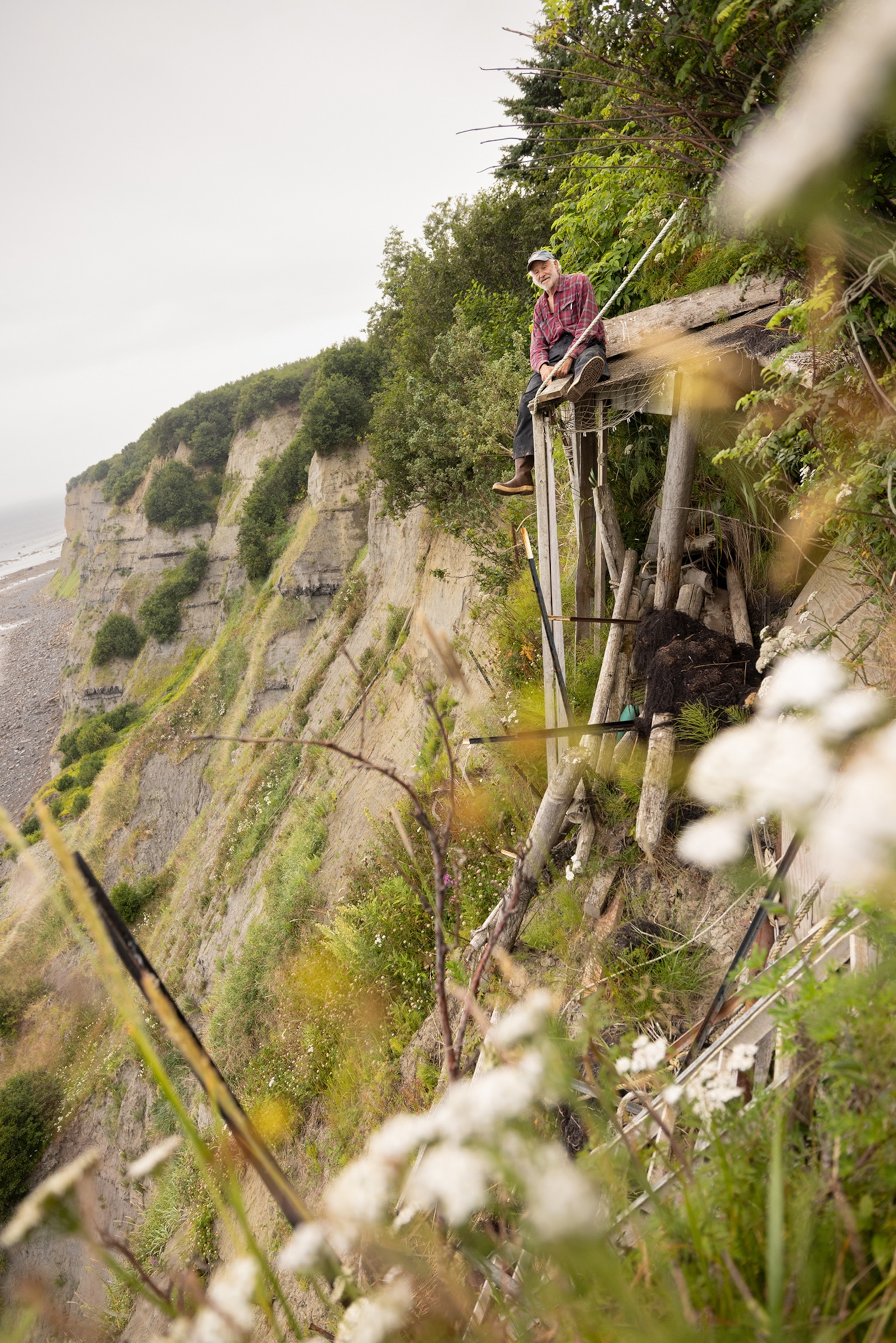 A man sits on the edge of a makeshift platform of mismatched pieces of wood, precariously built on the side of a high cliff.