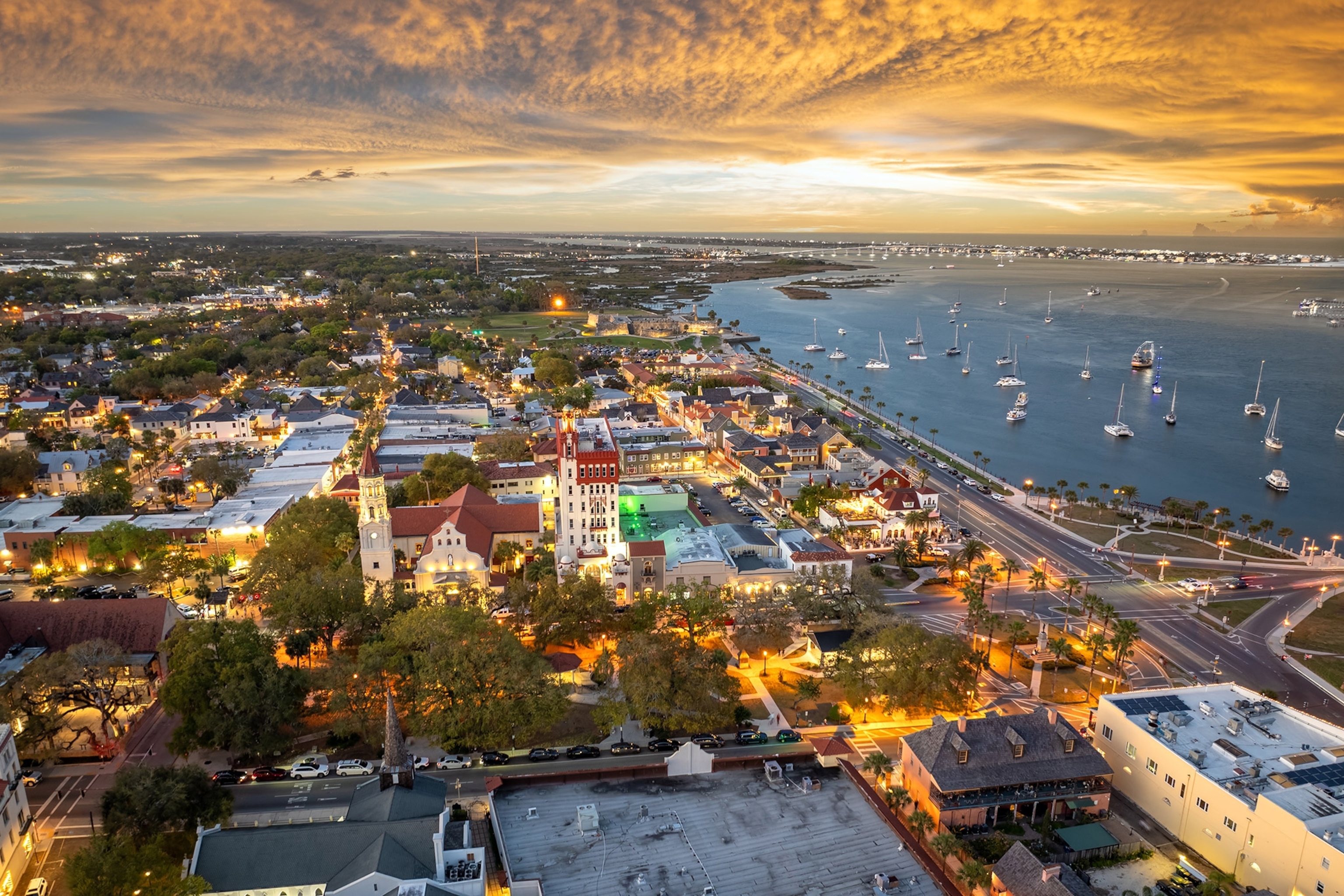 Aerial of a bay-side town glows with lights and under a dramatic sunset, with boats dotting the water beyond