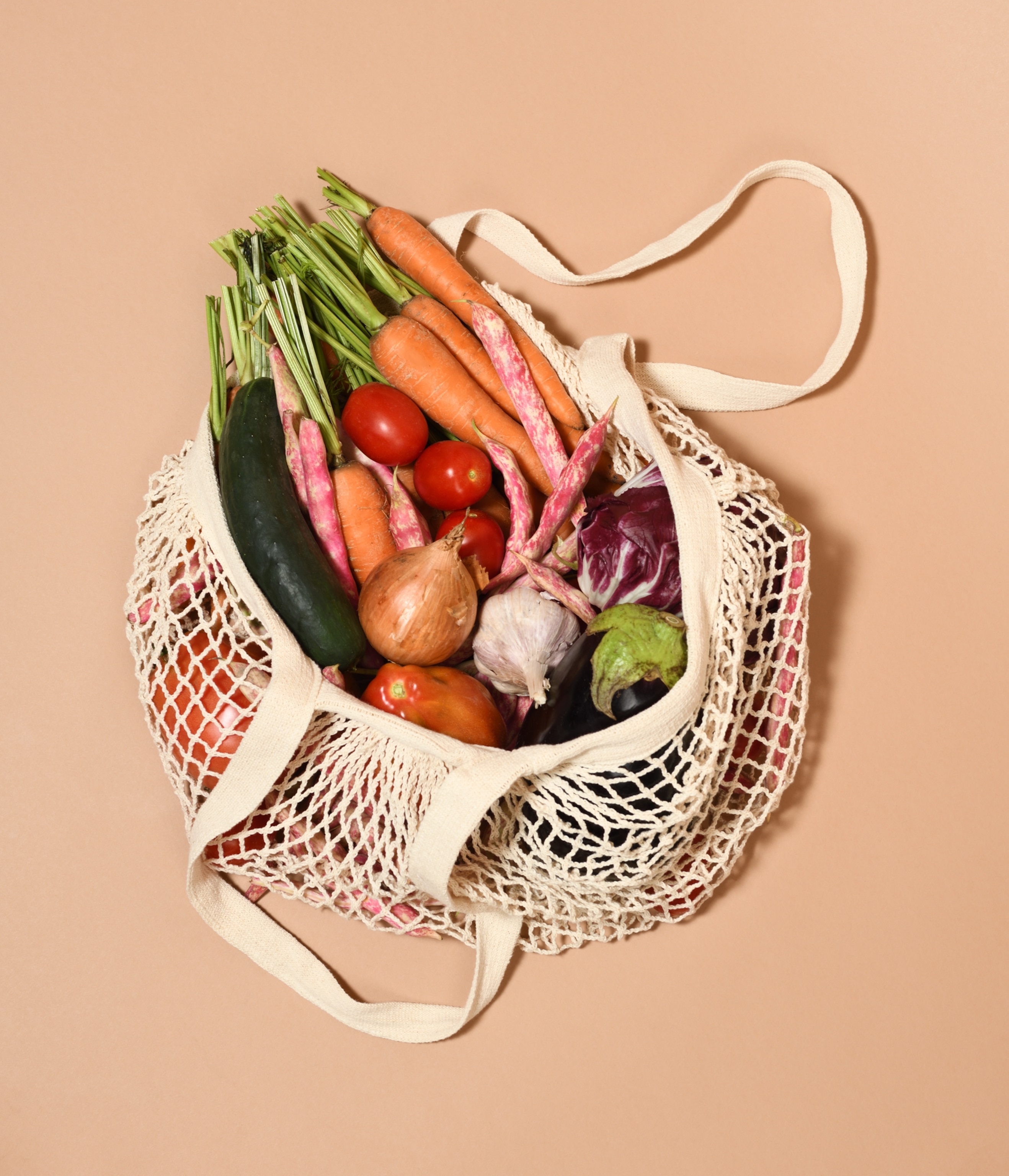 A beige, reusable net bag filled with vibrant vegetables, including carrots, tomatoes, onions, and lettuce, is placed on a light brown background.