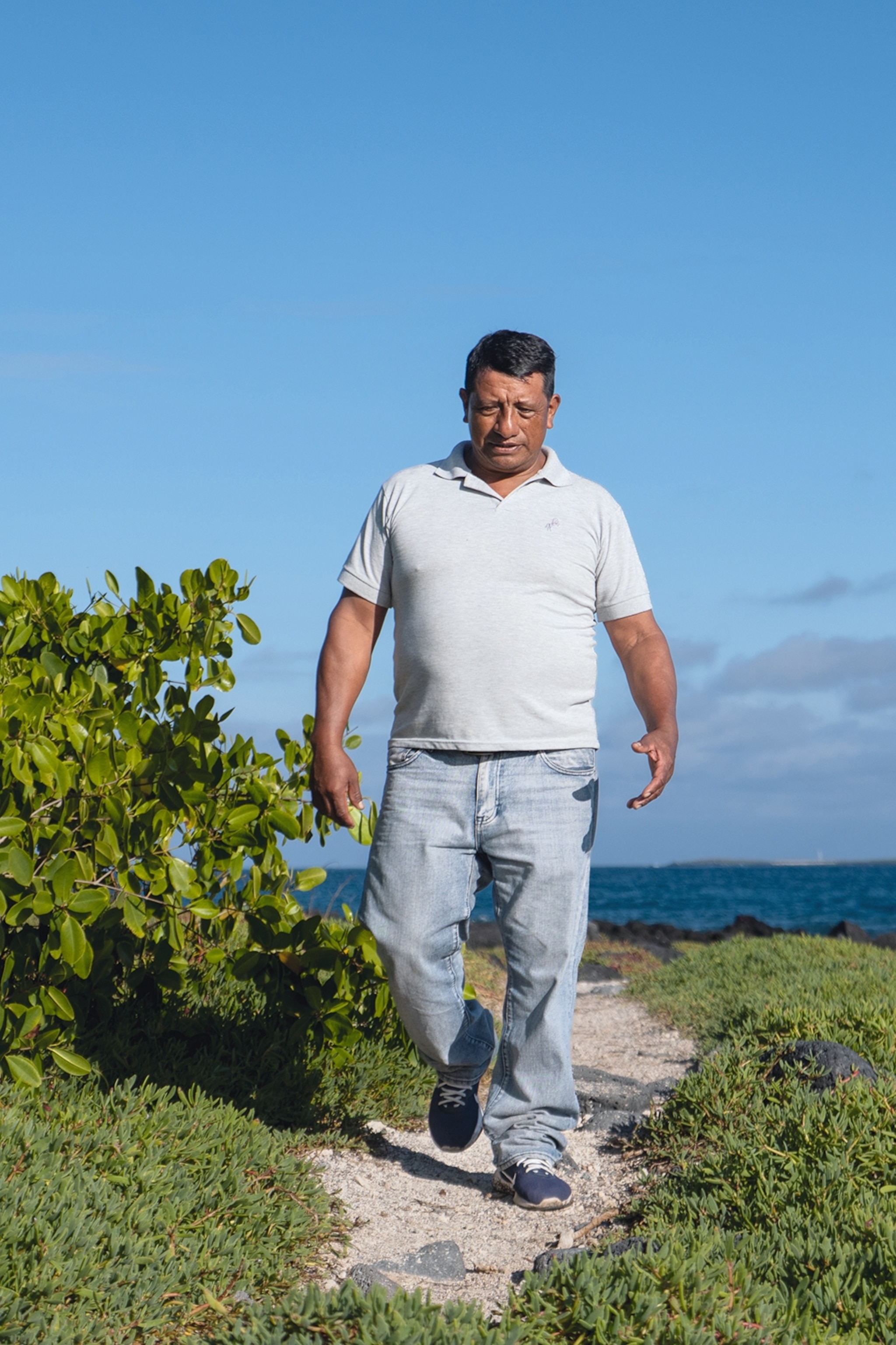 A middle-aged man in casual polo and jeans walking down a stony path on an island plateau with the ocean in the background.
