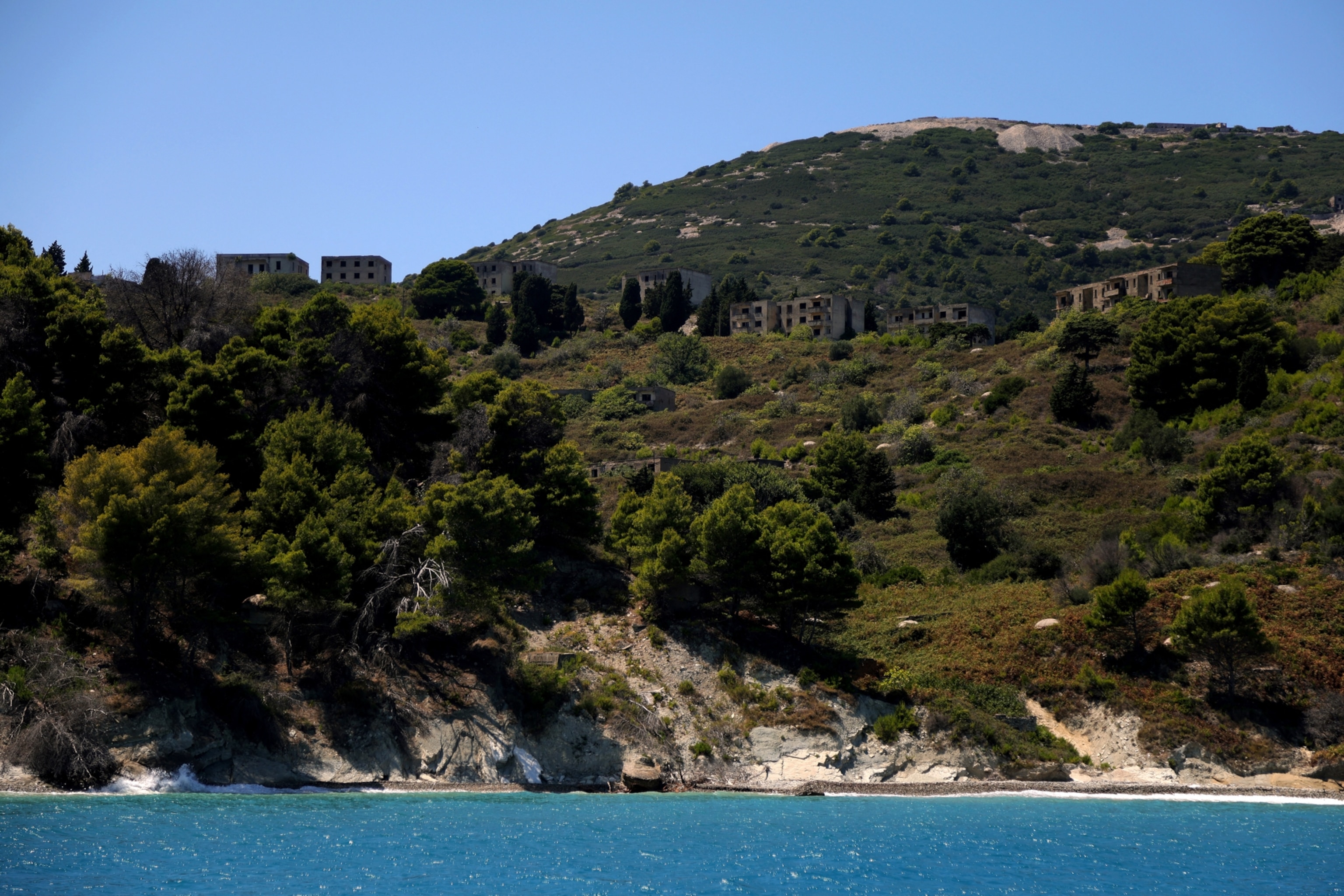 Albania's Sazar Island is seen from a distant view from the water, with various decaying buildings along the landscape of trees.