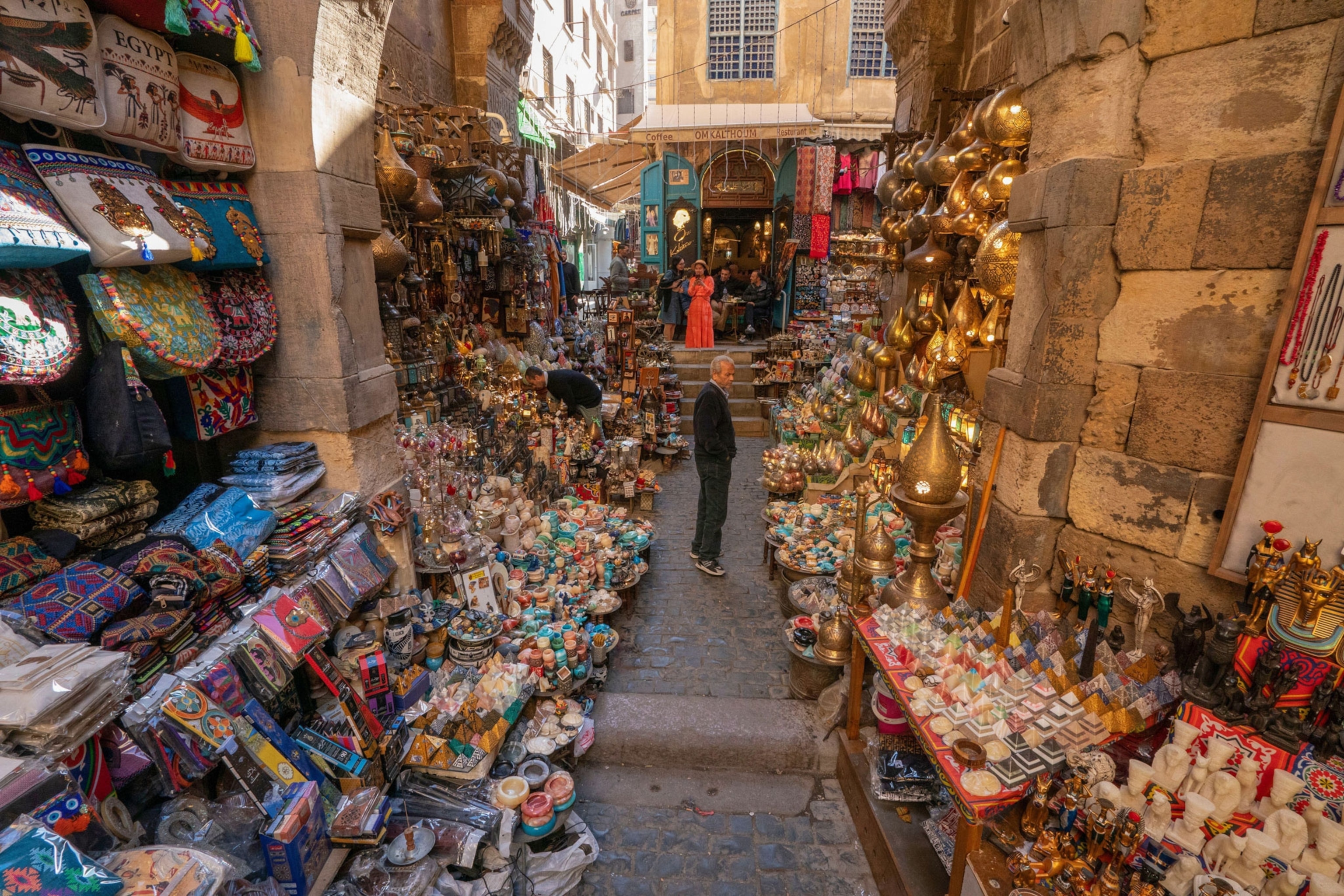 A man standing in the Khan Al-Khalili market in Cairo, surrounded by Egyptian goods in an old brick and cobblestone corridor.