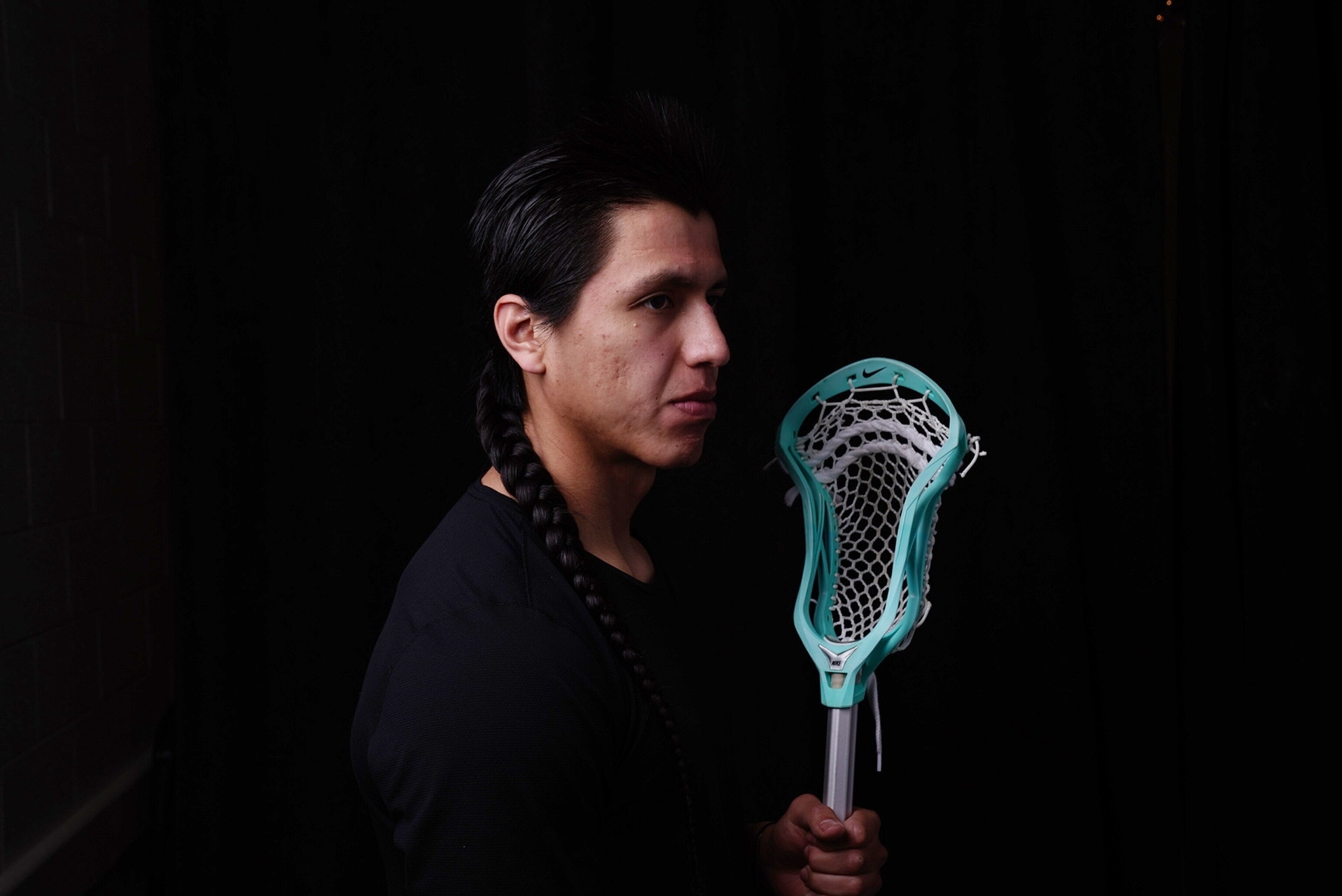 Lacrosse player Lyle Thompson in profile against a black background holding a blue lacrosse stick