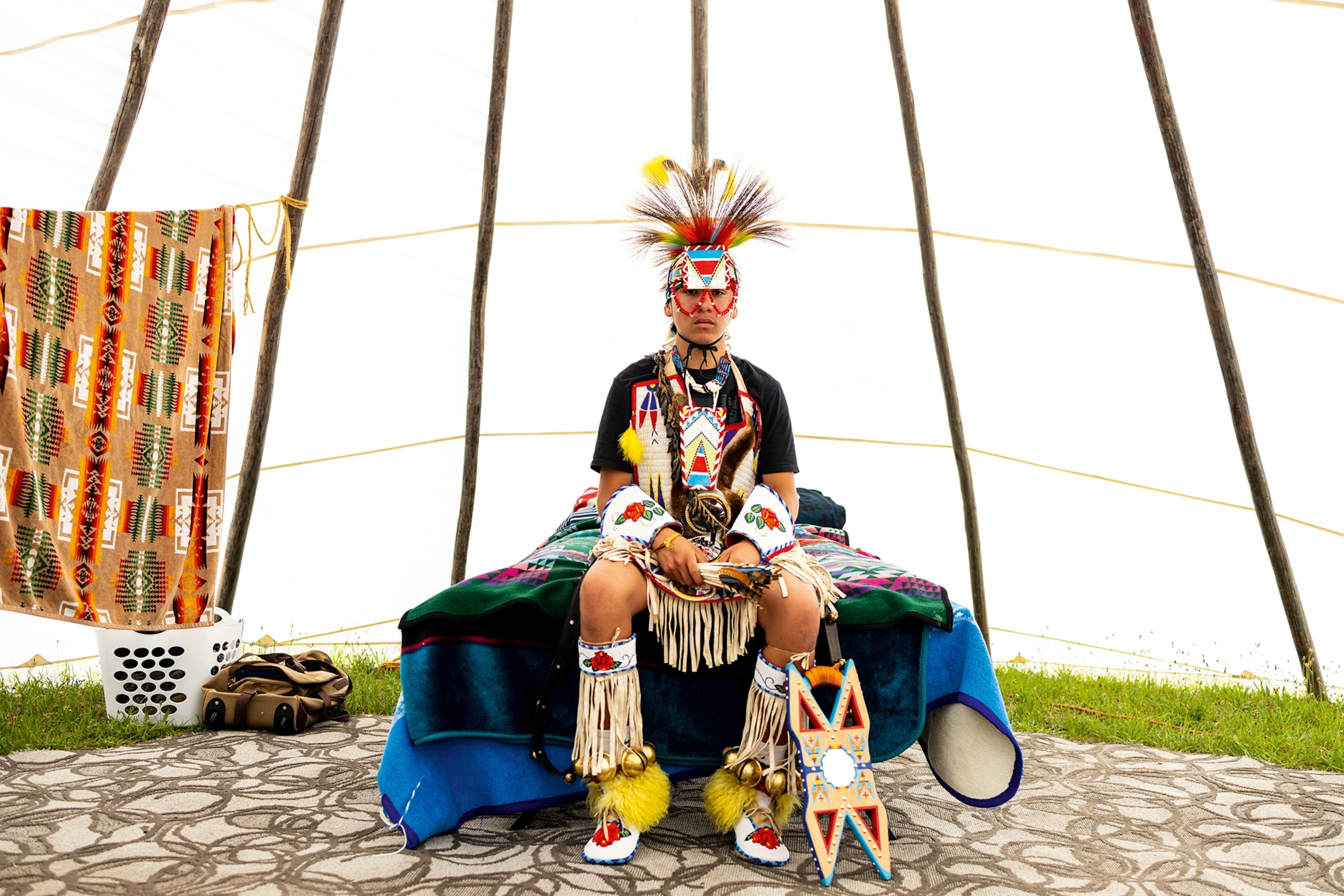 A young Native American man sitting on the end of his camping bed inside a tipi, dressed in festive clothes with a feathered head dress and tassels around his hips and calves.