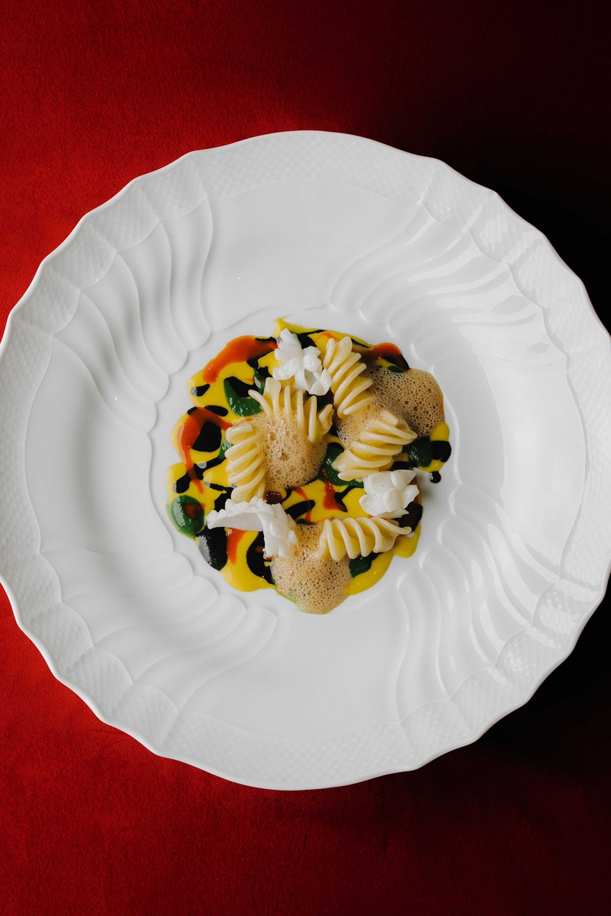 A pasta dish sits on a white plate on top of a dark red tablecloth.