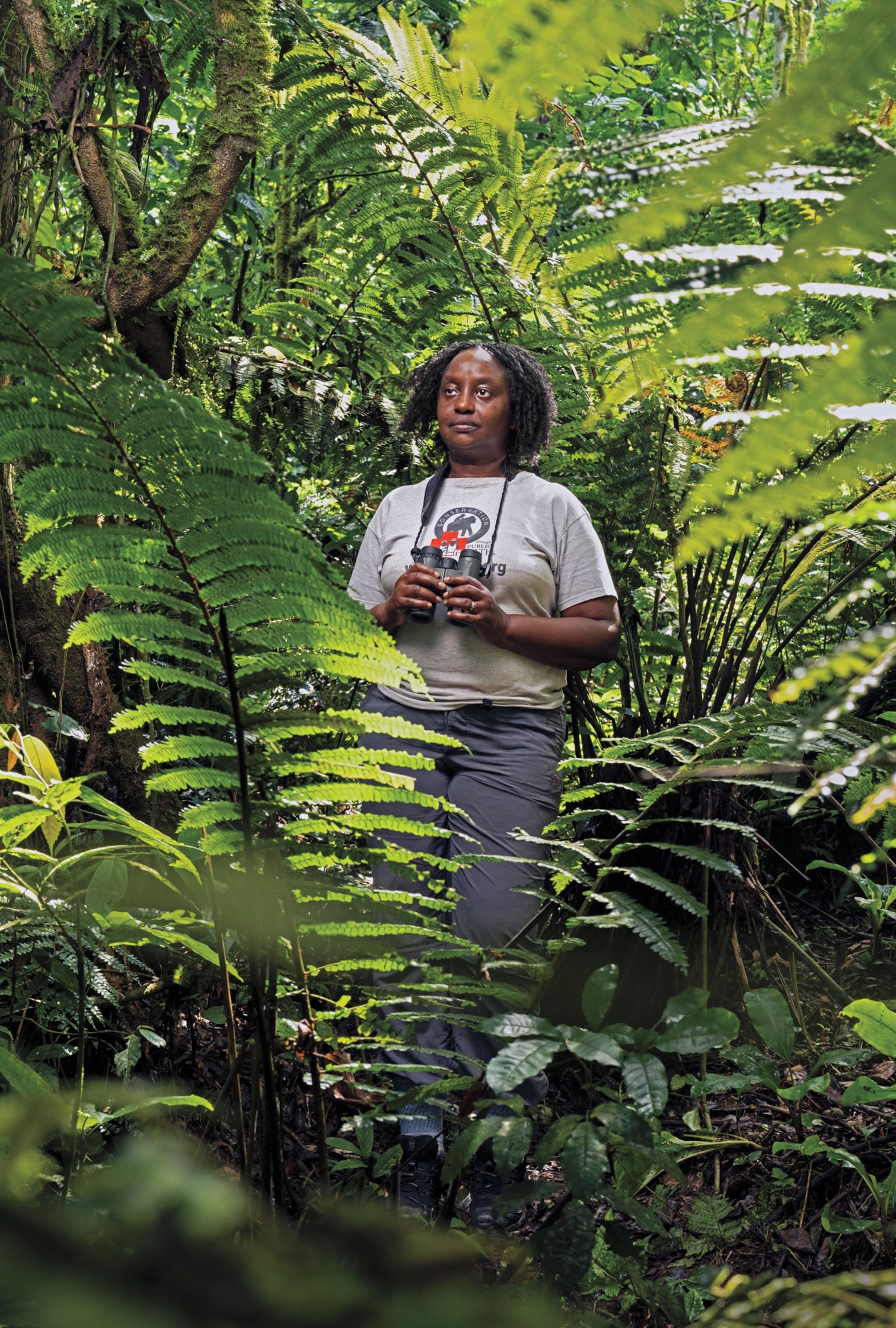 Portrait of a woman wearing grey pants and a grey shirt looks off to the distance while her hands hold binoculars hanging around her neck. She is standing between trees in a forest.