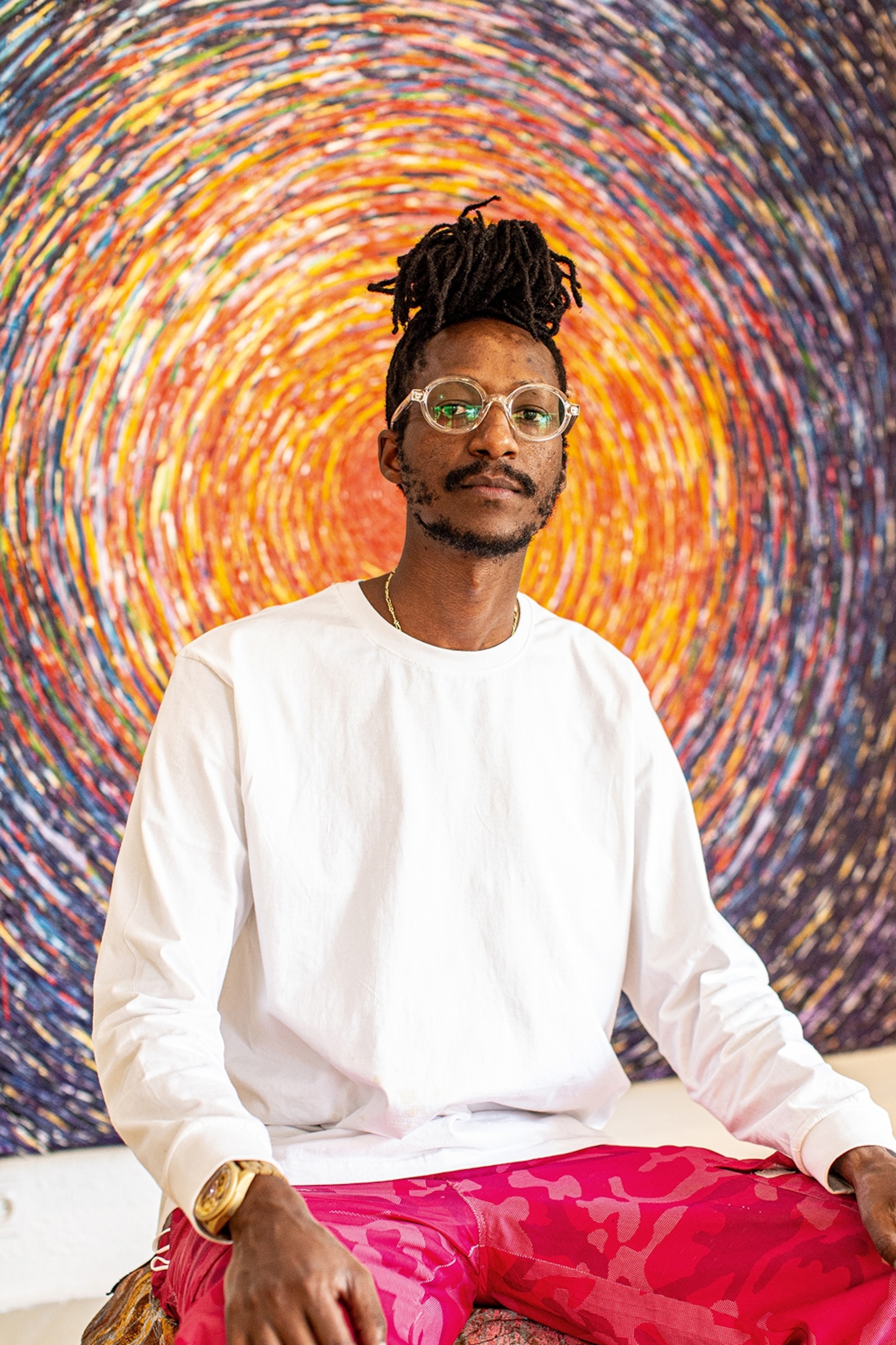 A local artist with dreadlocks in a bun and glasses, posing in front of a painting.