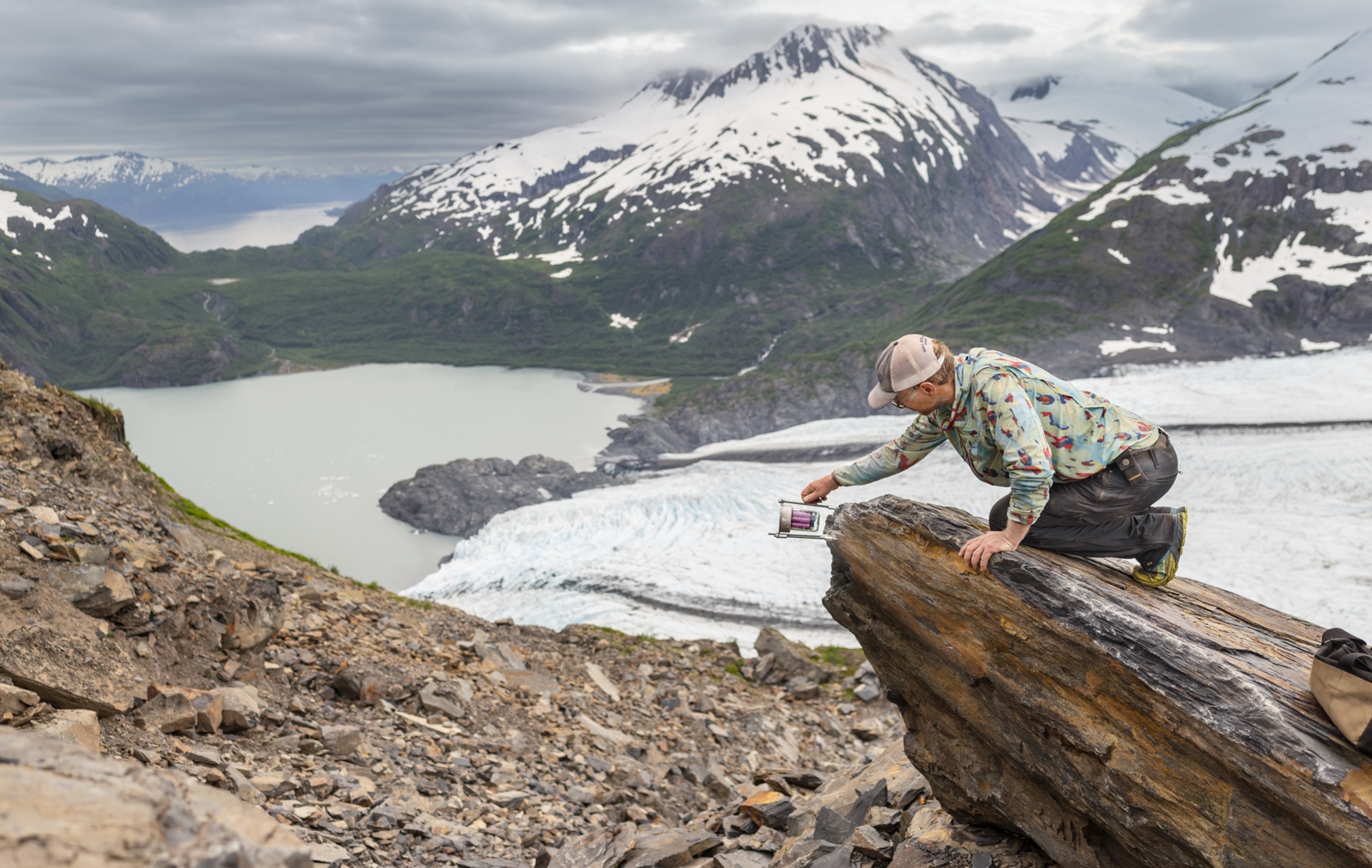 A person kneels on a large rock near a glacier and a pale lake, holding an instrument, with snowy mountains and green hills in the background.