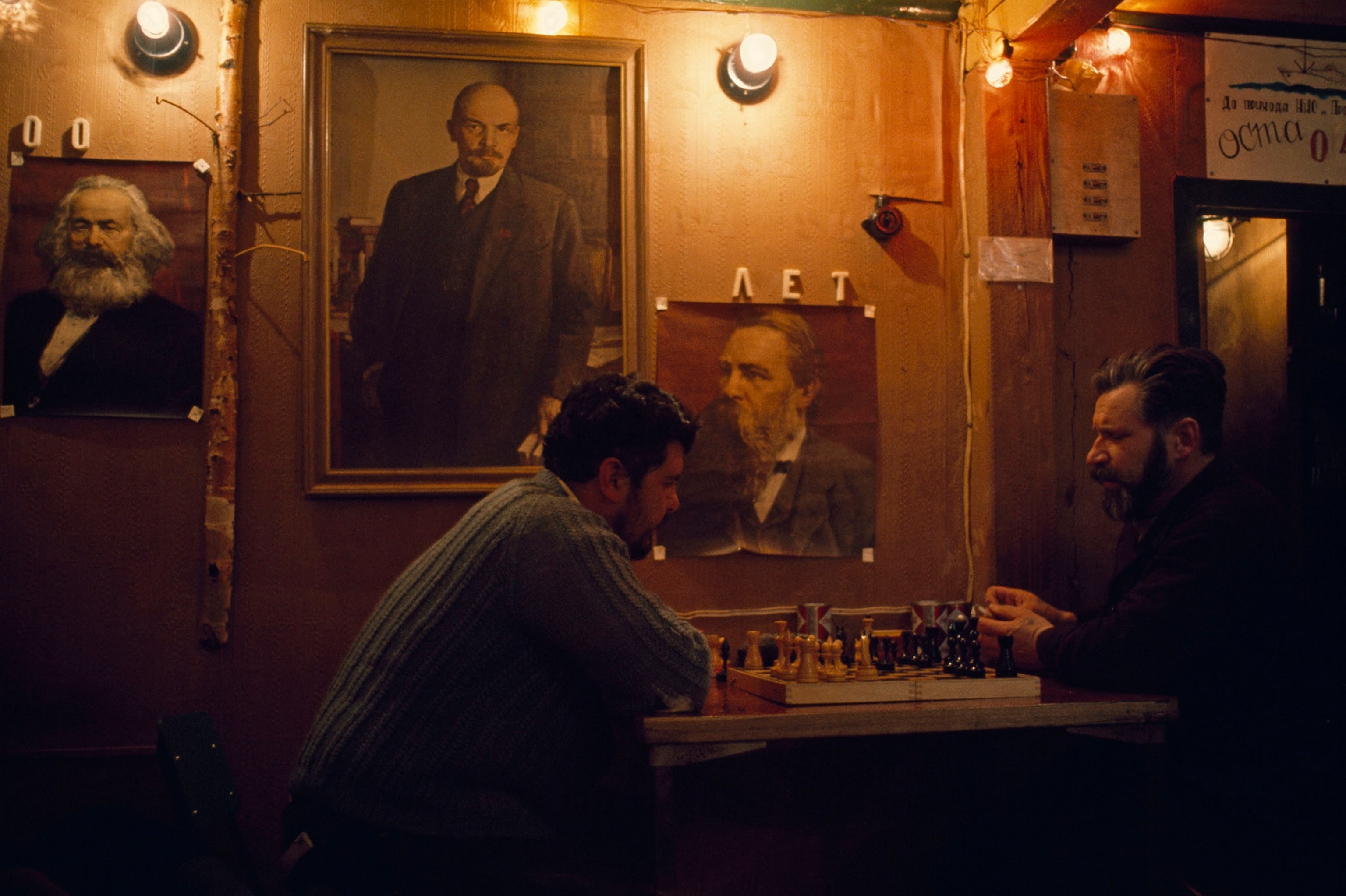 Two Russian scientists play chess beneath portraits of famous people.