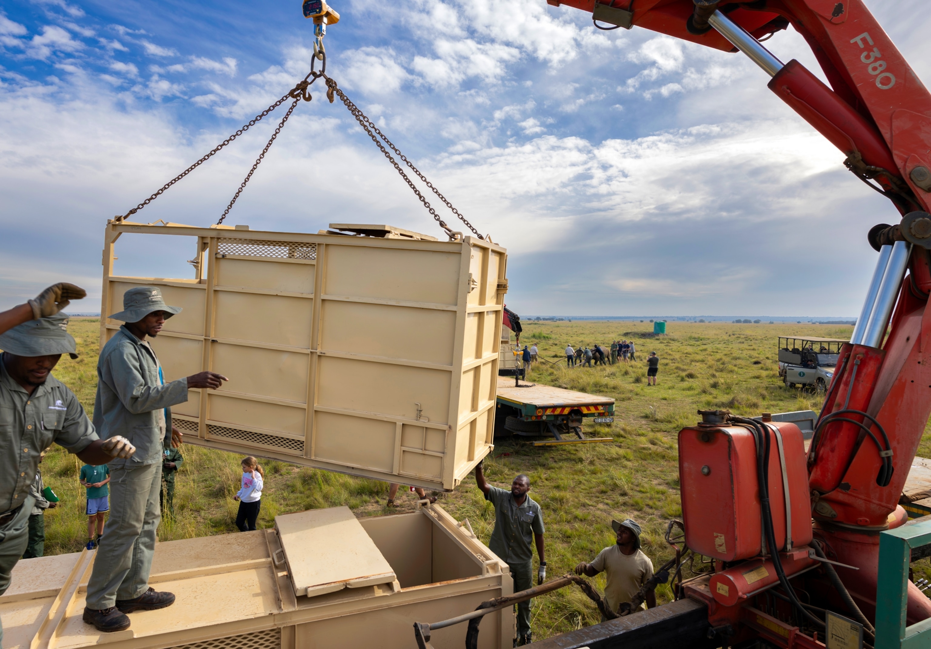 Two men stand on top of a large metal container while two men stand on the ground to the right of them. They assist in directing a large red crane that is lifting a different metal shipping container.
