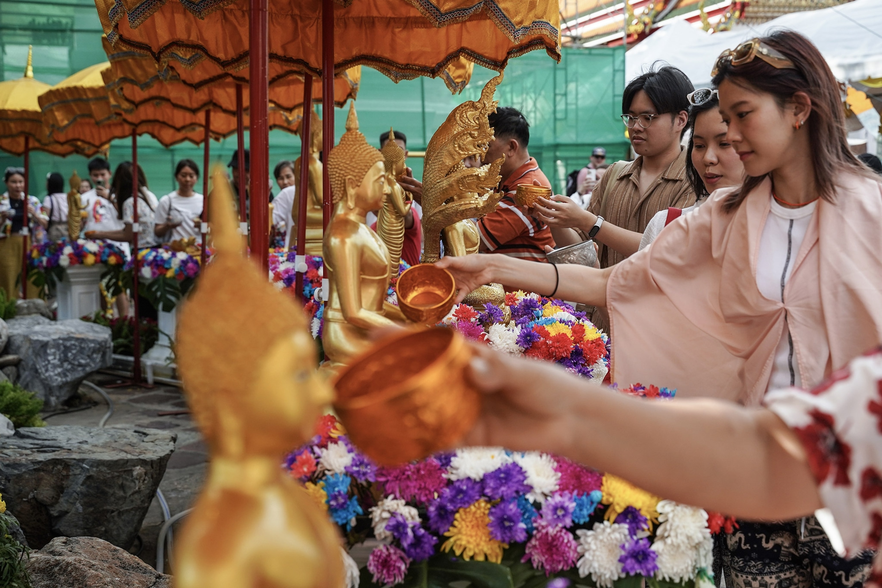 People crowd around a sheltered table laden with gold Buddha statues, pouring water from copper bowls, flowers strewn about.