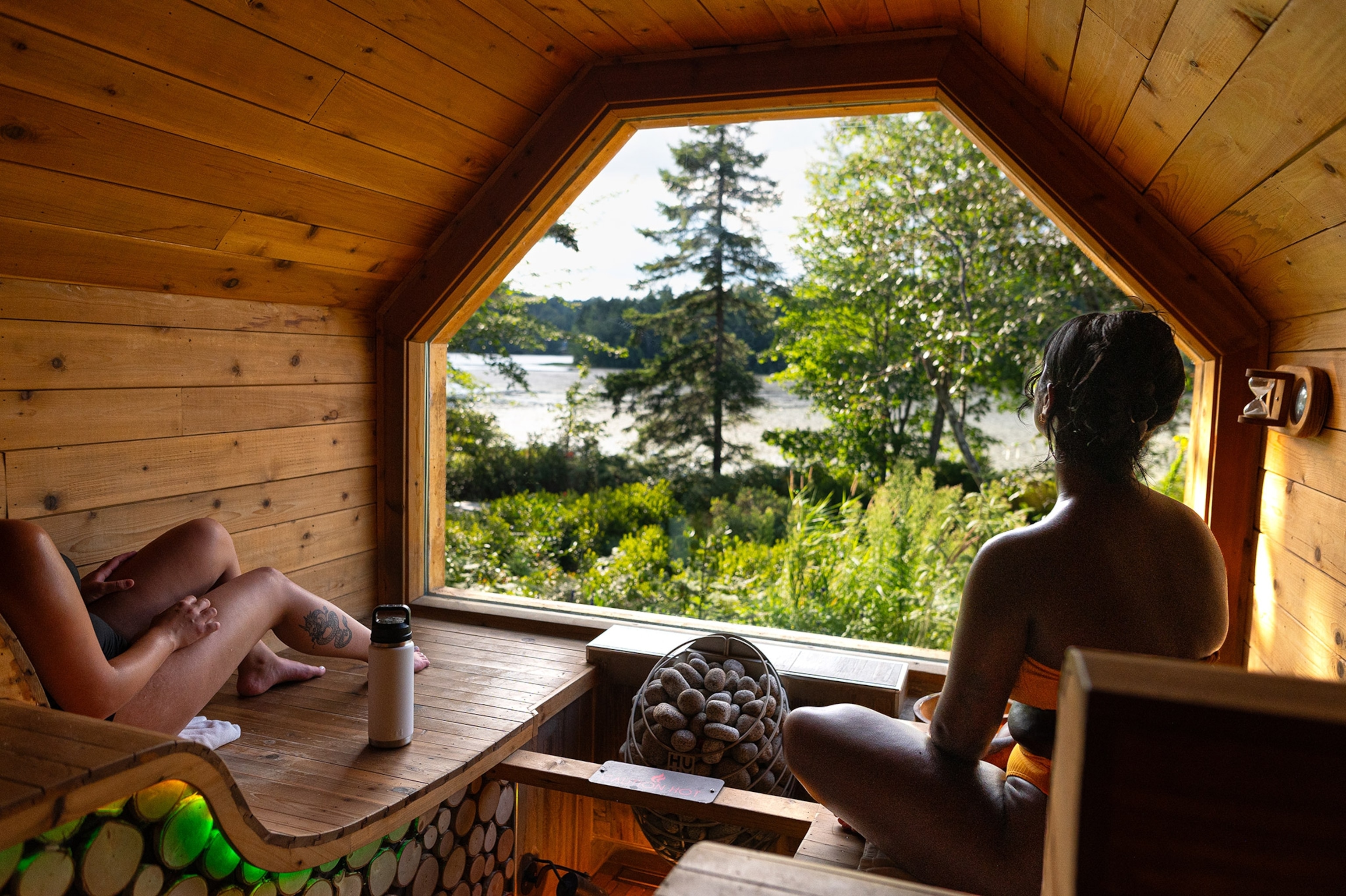 Two women sit on lounges in a small wood-paneled space, facing a large window looking out into wooded natural area