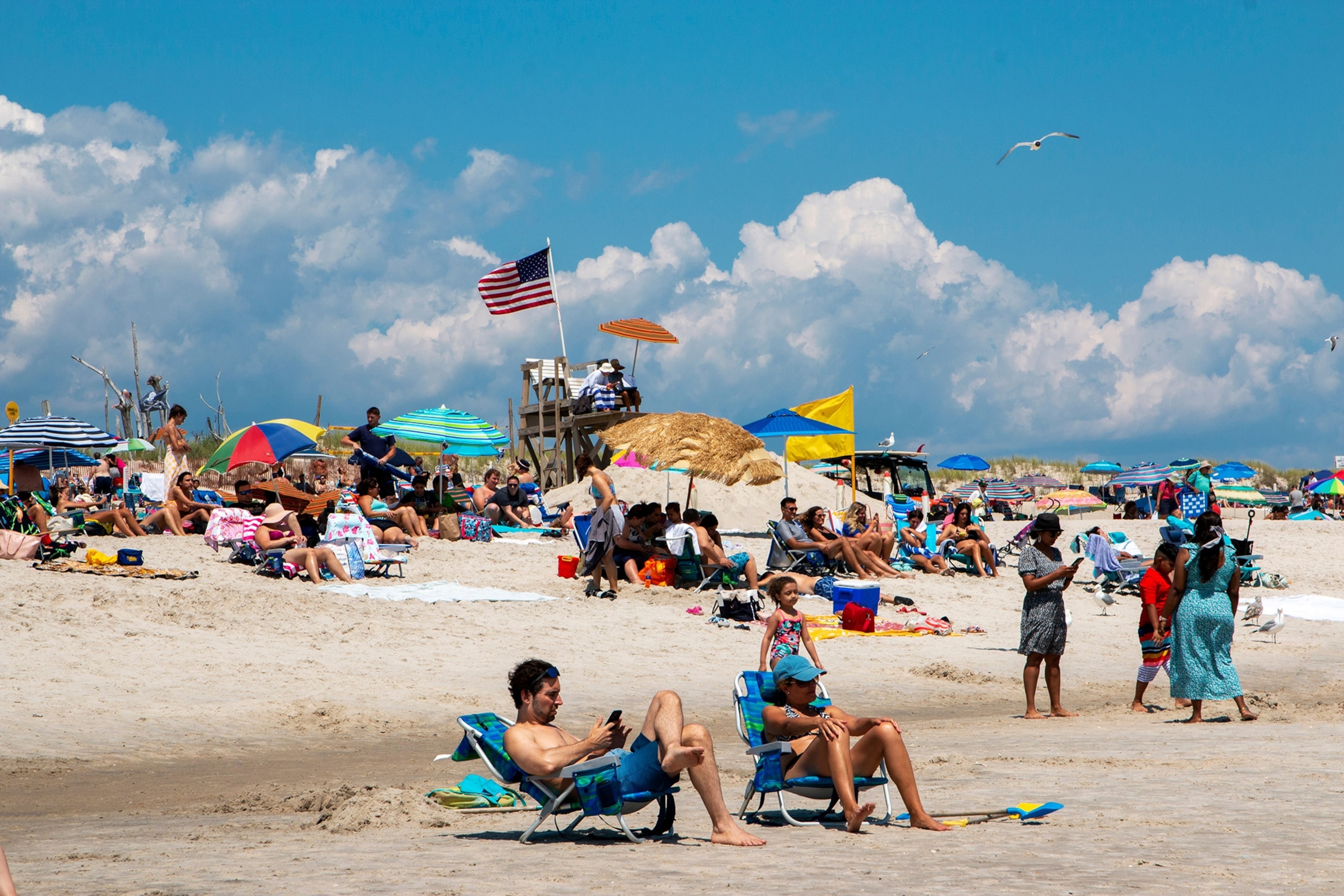 A group of beachgoers on a beach beneath a cloudy sky, a lifeguard tower and umbrellas, an American flag flying in the background.