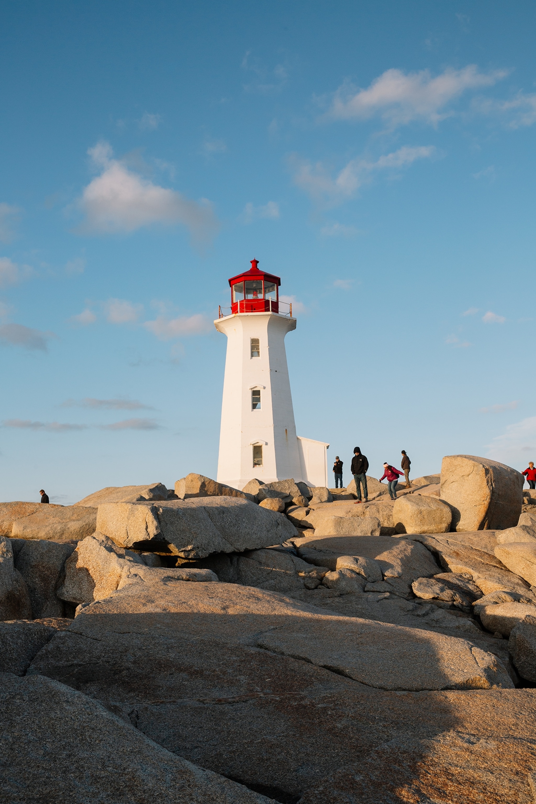 A white lighthouse with a red lantern room stands on a rocky shoreline, while several people walk among large granite boulders under a clear blue sky.