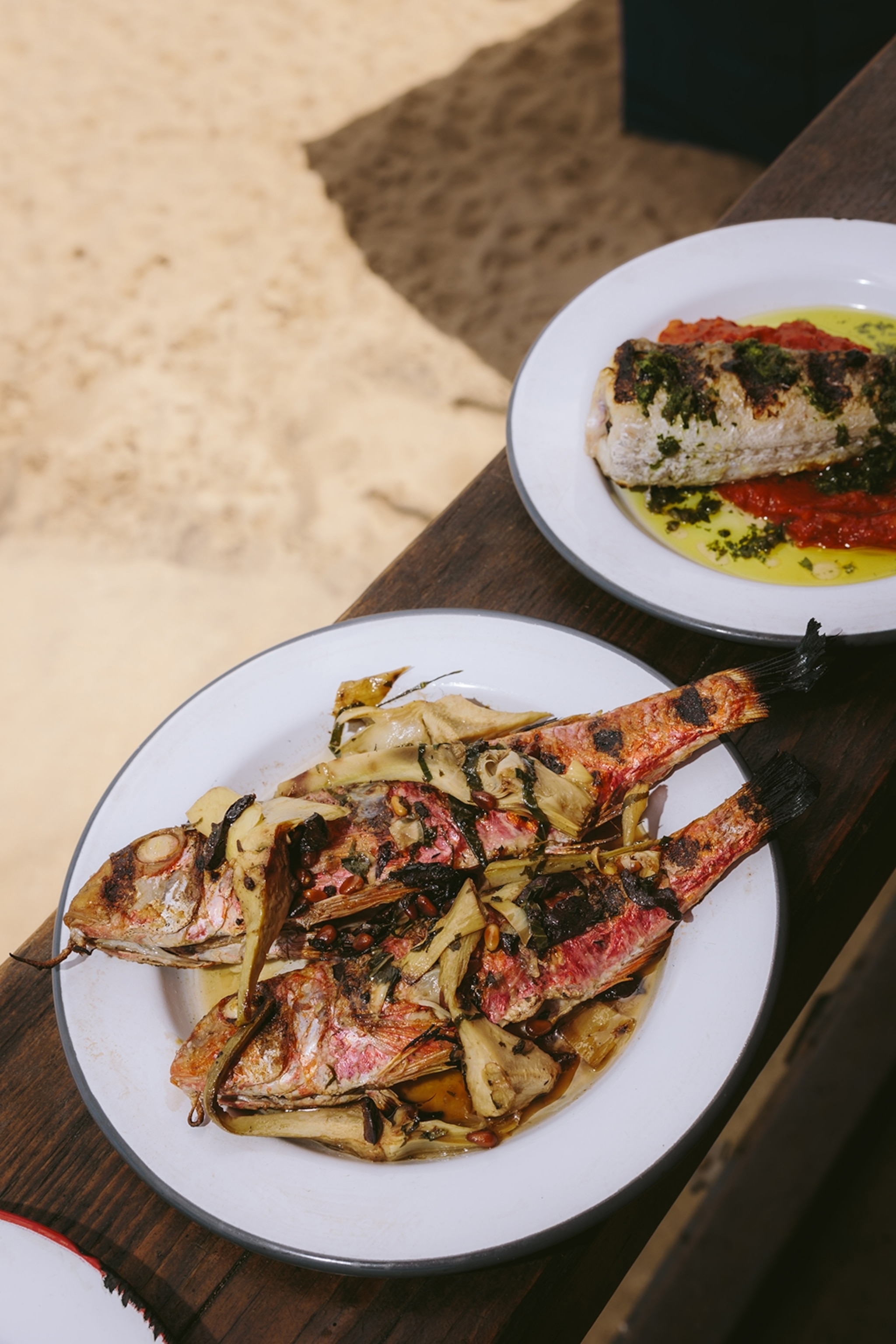 Two plates of grilled fish dishes with vibrant sauces on a wooden railing with a beach below.