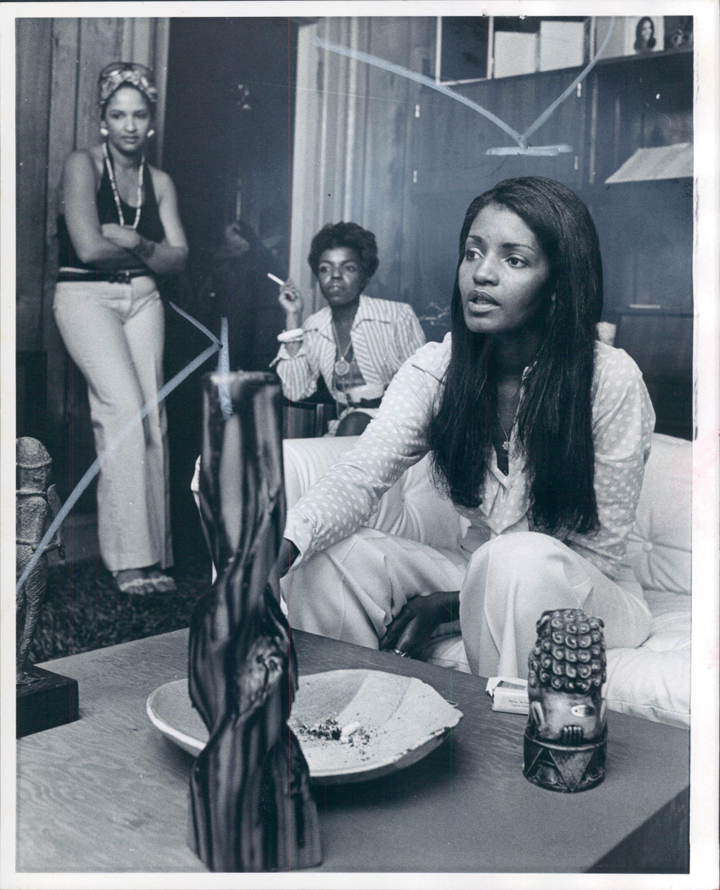 A black and white image of three women in a living room. One sits on a sofa, engaged, while the others stand behind, relaxed