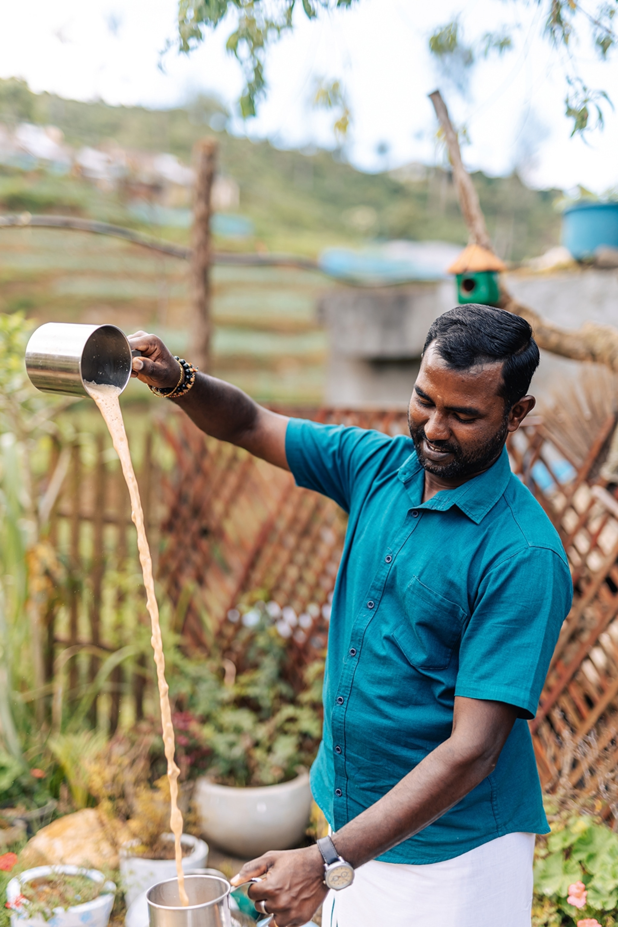 A local man pouring a liquid from a height into a tin cup with a simple garden in the background.
