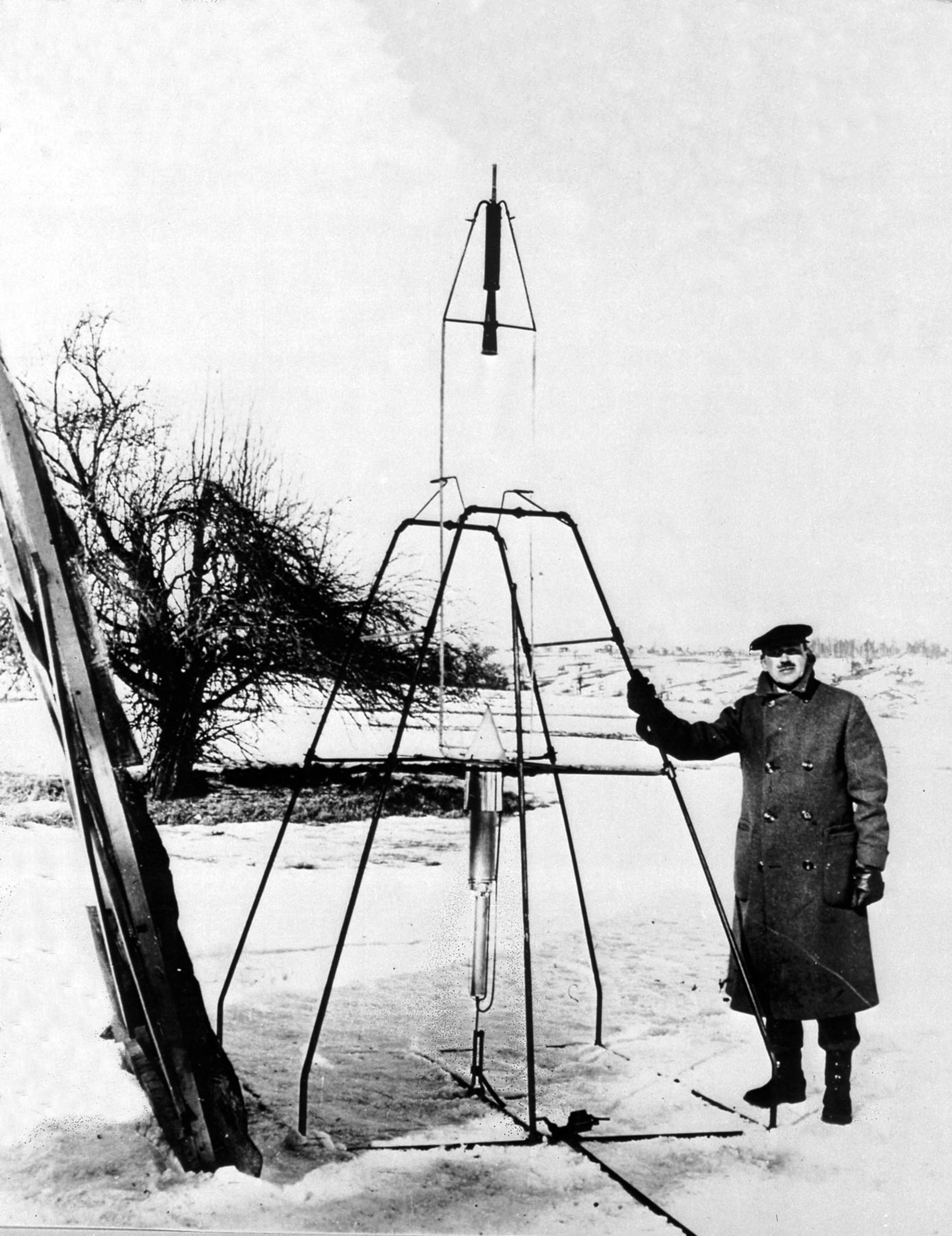 A man in a heavy coat stands beside an early rocket model set on a snowy field. Behind him, leafless trees and distant hills are visible