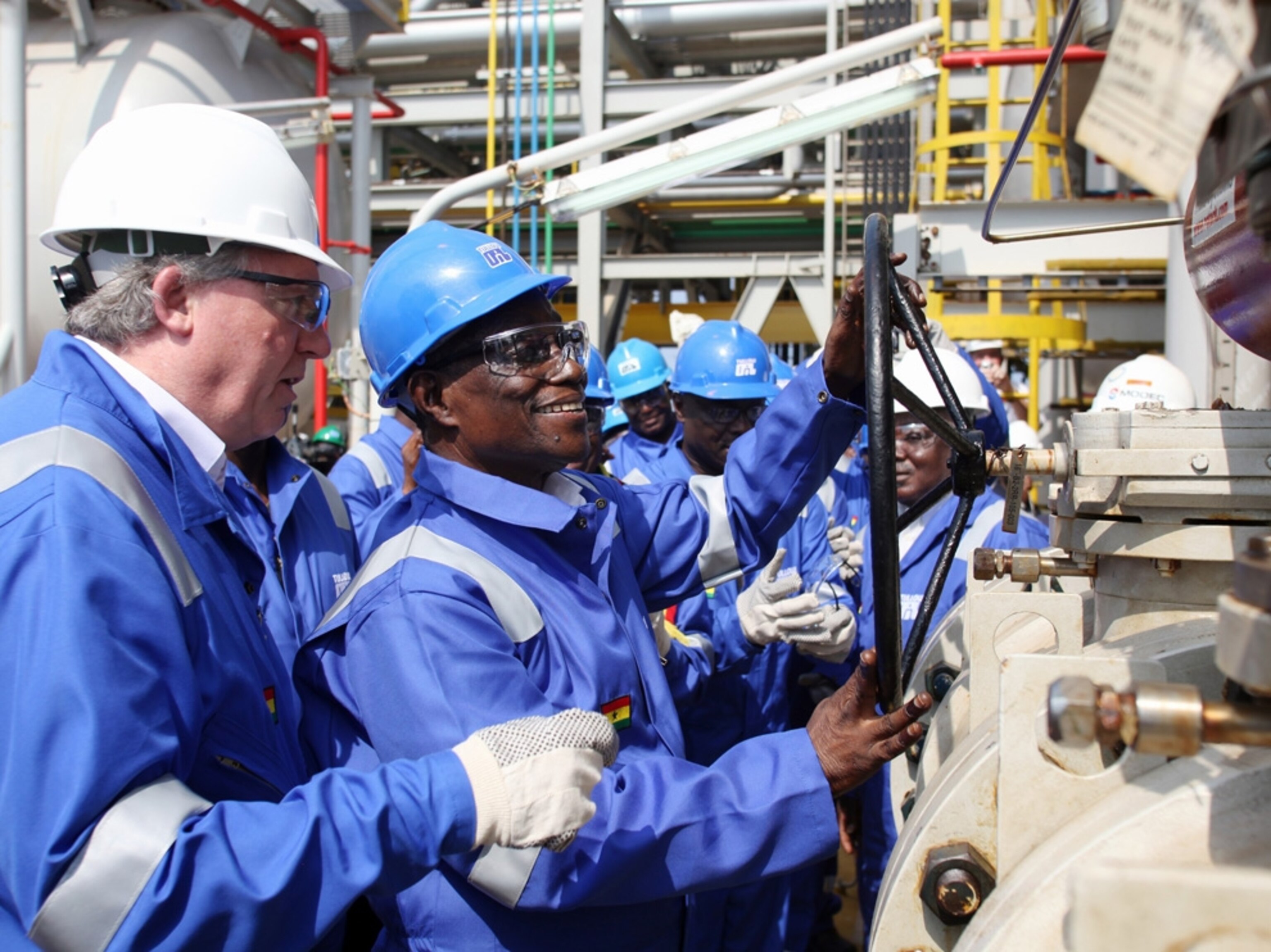 Men in blue coveralls and hard hats watch Ghana’s president turn the valve to officially open an oil rig