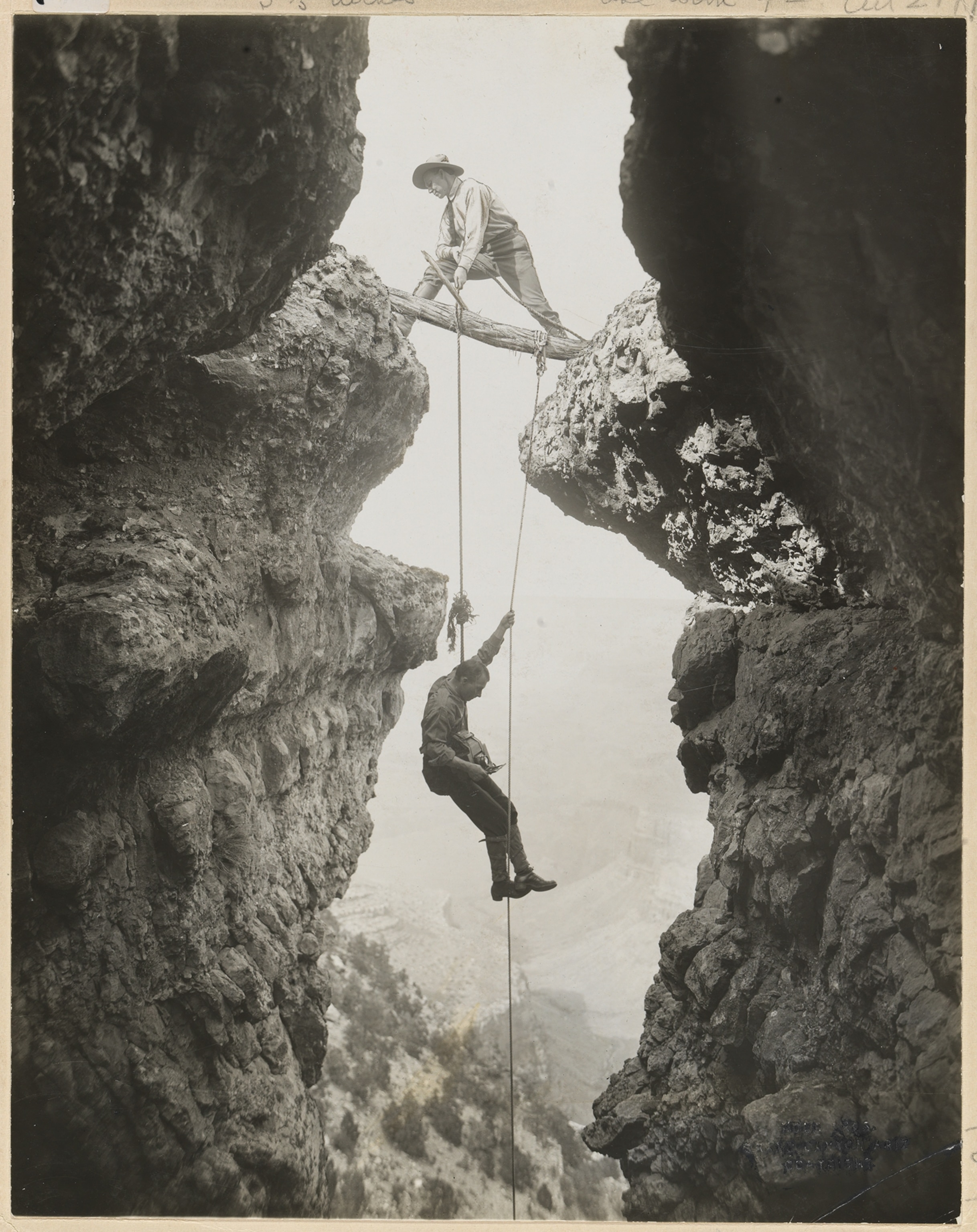 A man stands on a branch over an abyss, lowering another man into the Grand Canyon by rope