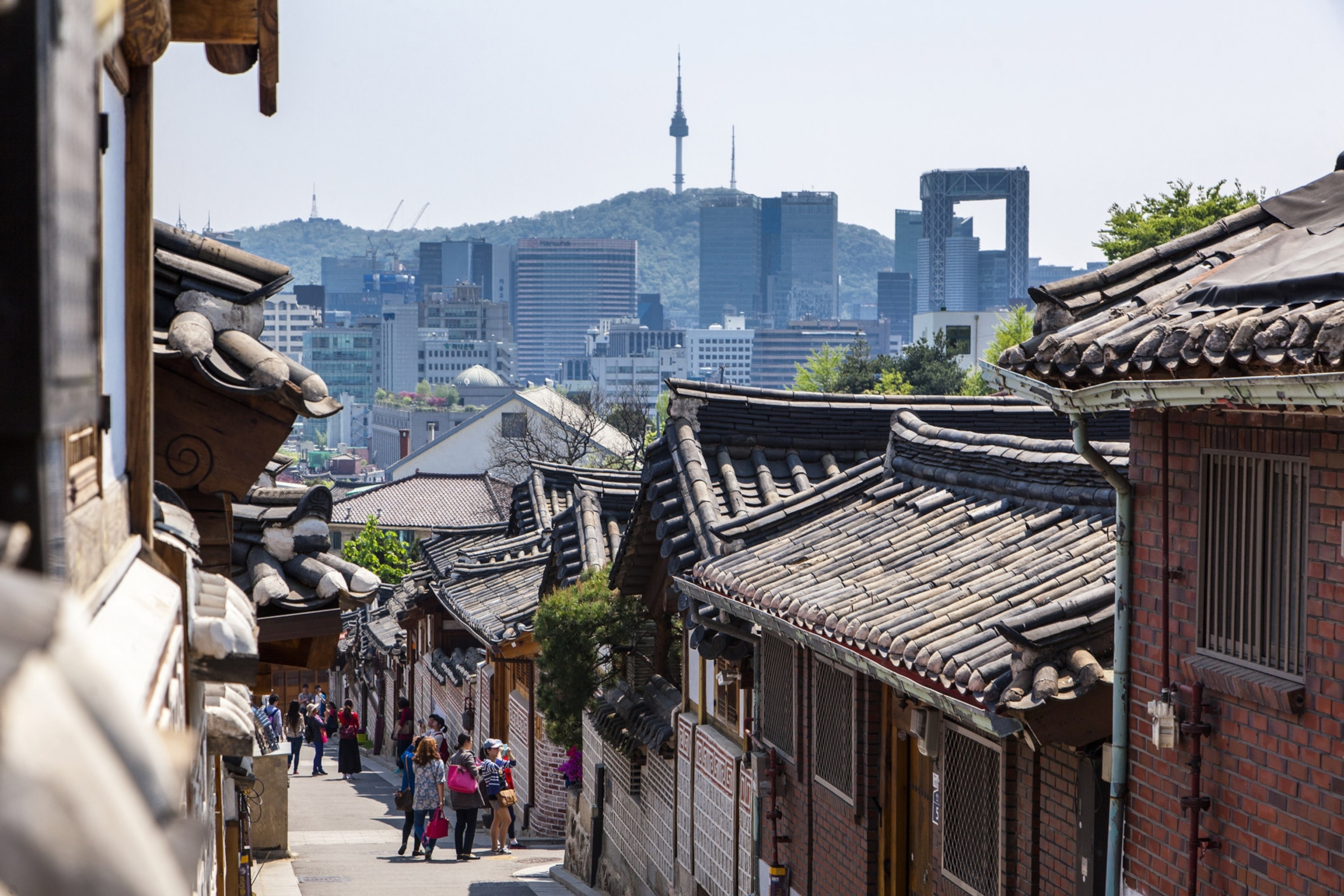 A narrow street surrounded by traditional architecture, people visible at the bottom of a hill, city skyline in the distance