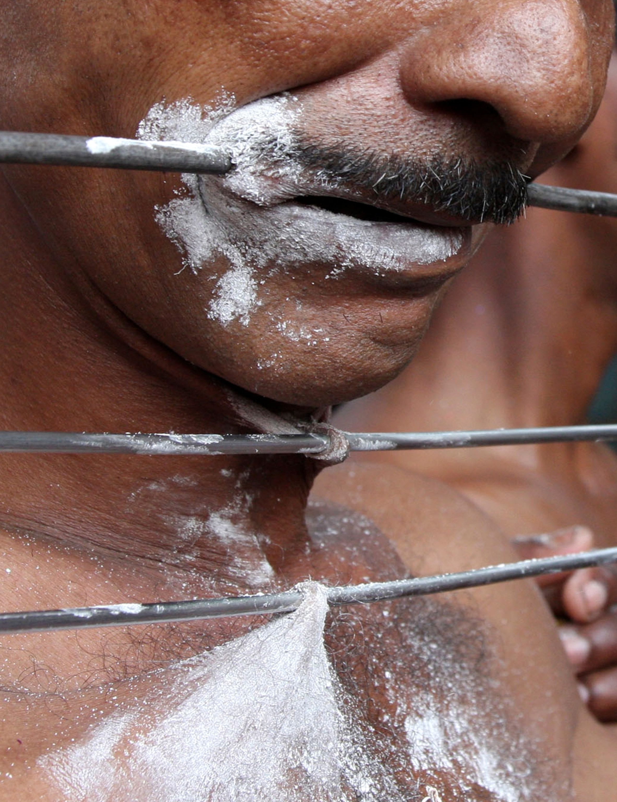 A close-up view of a man's face and neck, where metal skewers are seen pierced through the skin of his upper lip, neck, and chest