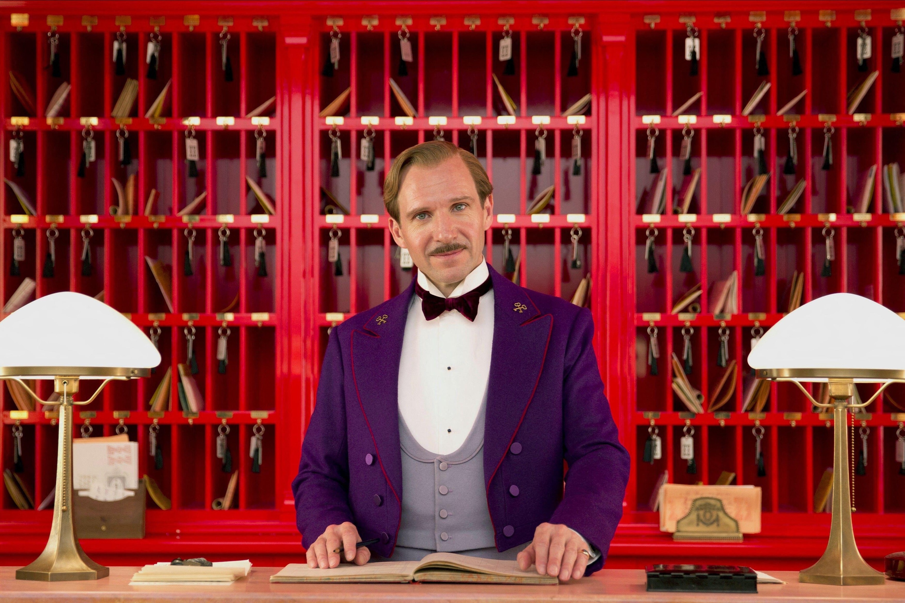 Ralph Fiennes in the role of concierge Gustave H from The Grand Budapest Hotel behind a reception counter.