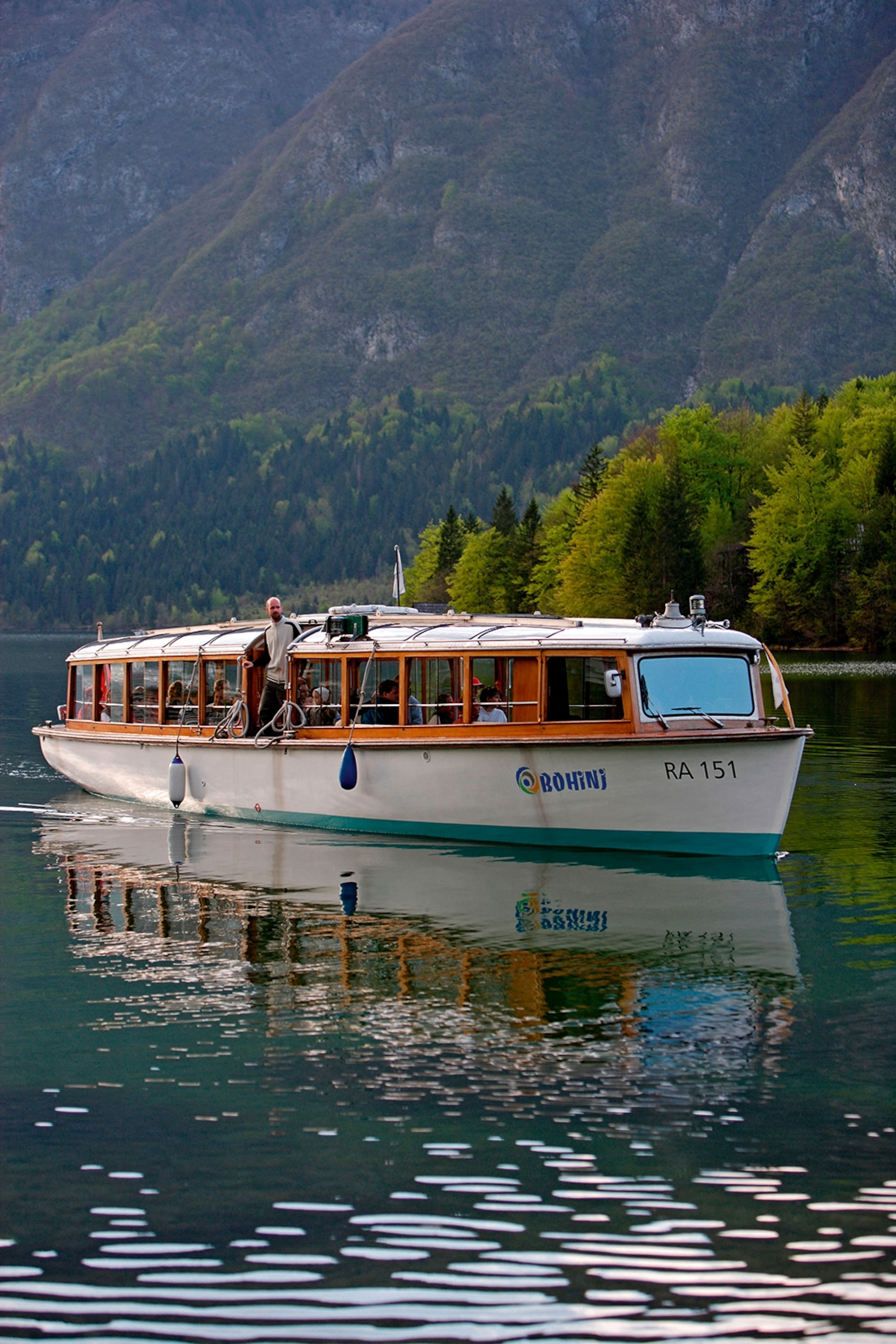 A small lake cruise ship gliding over clear water with trees and hill in the distance.