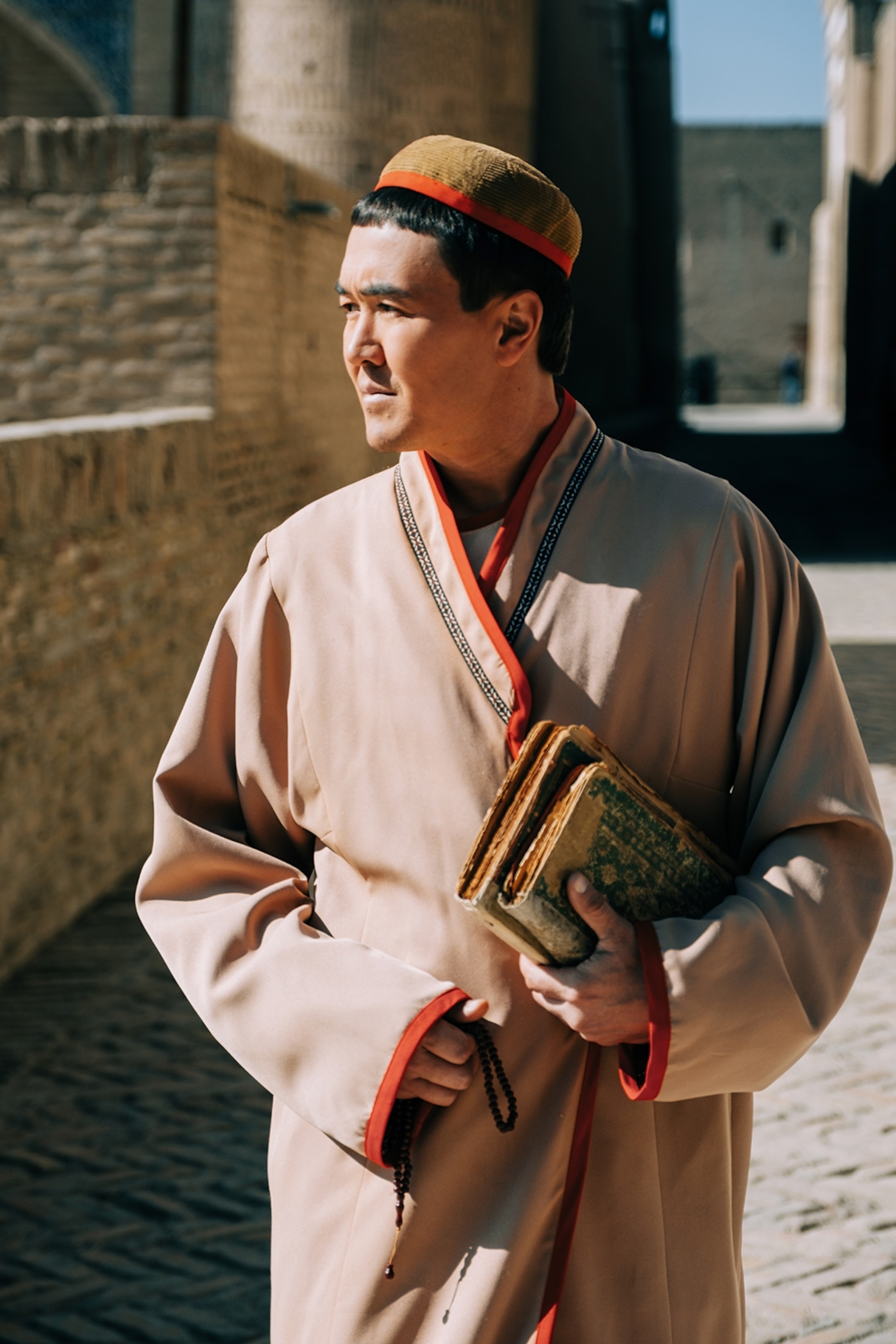 A portrait of a local man dressed in a silk tunic and rounded hat on the crown of his head, walking through the cobble-stoned streets of Khiva, carrying old books.