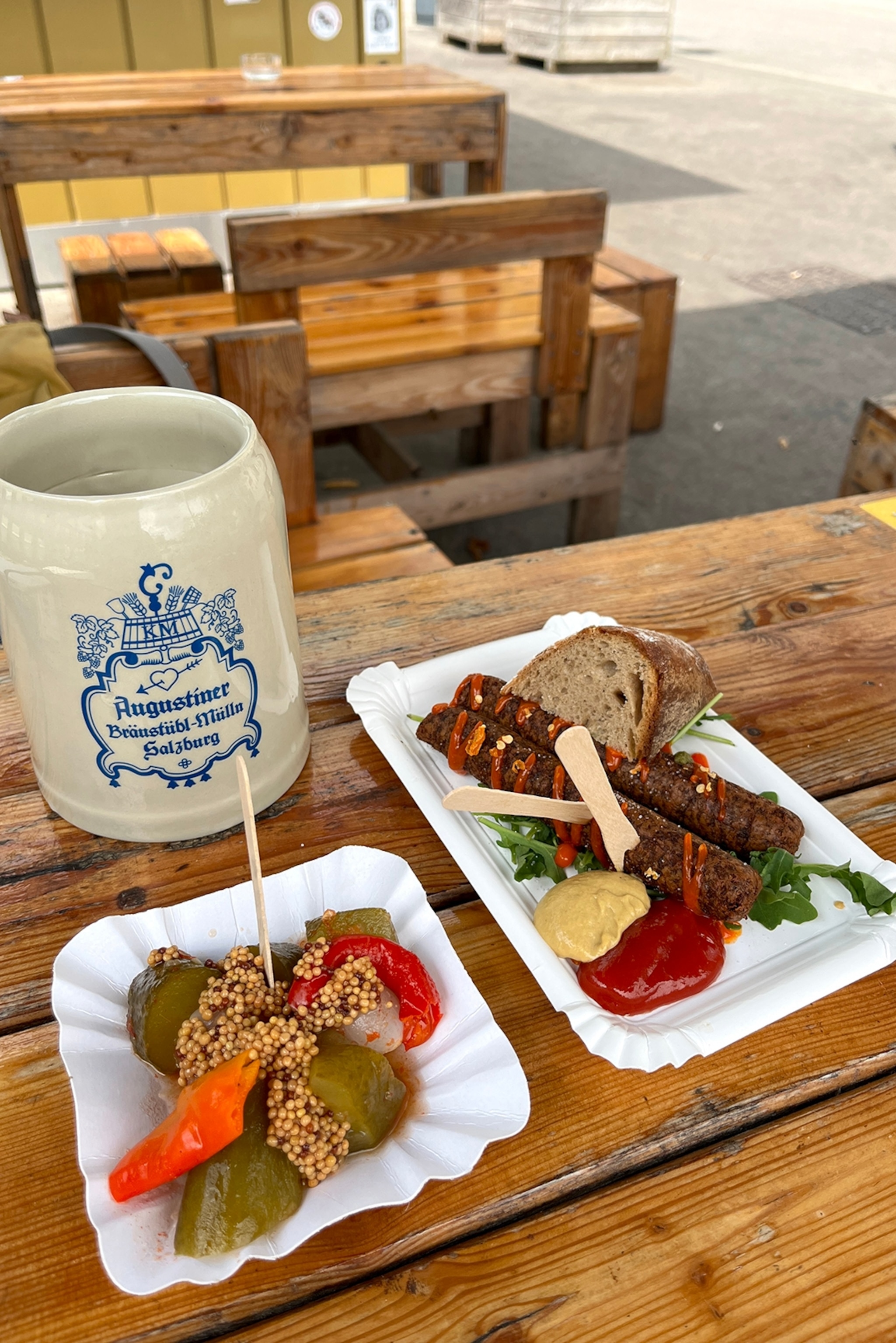 A casual image of two paper plates on a rustic outdoor table with sausages, pickles, condiments and a traditional beer mug.