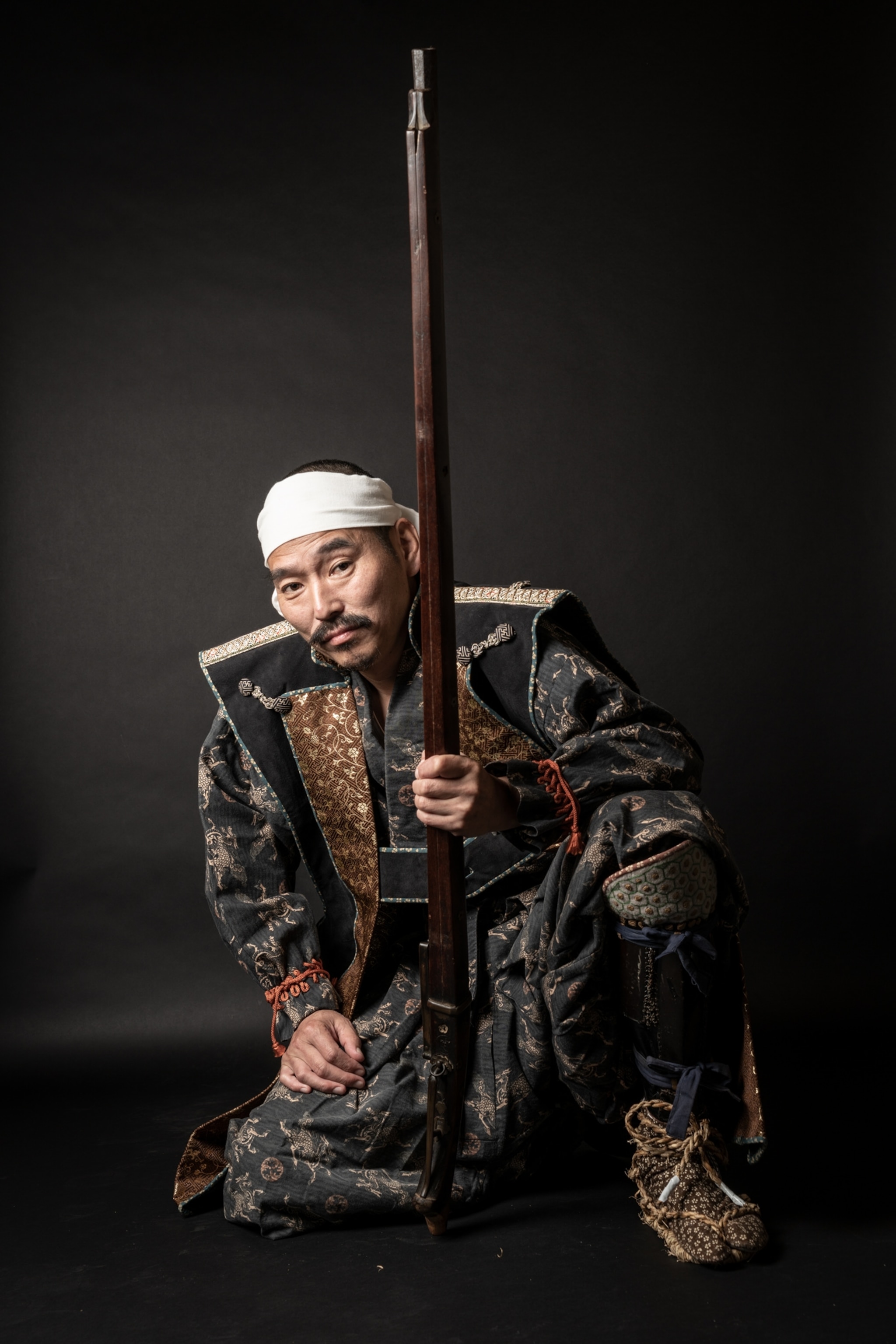 Picture of man with long long mustache and beard in white headband standing on his right knee.