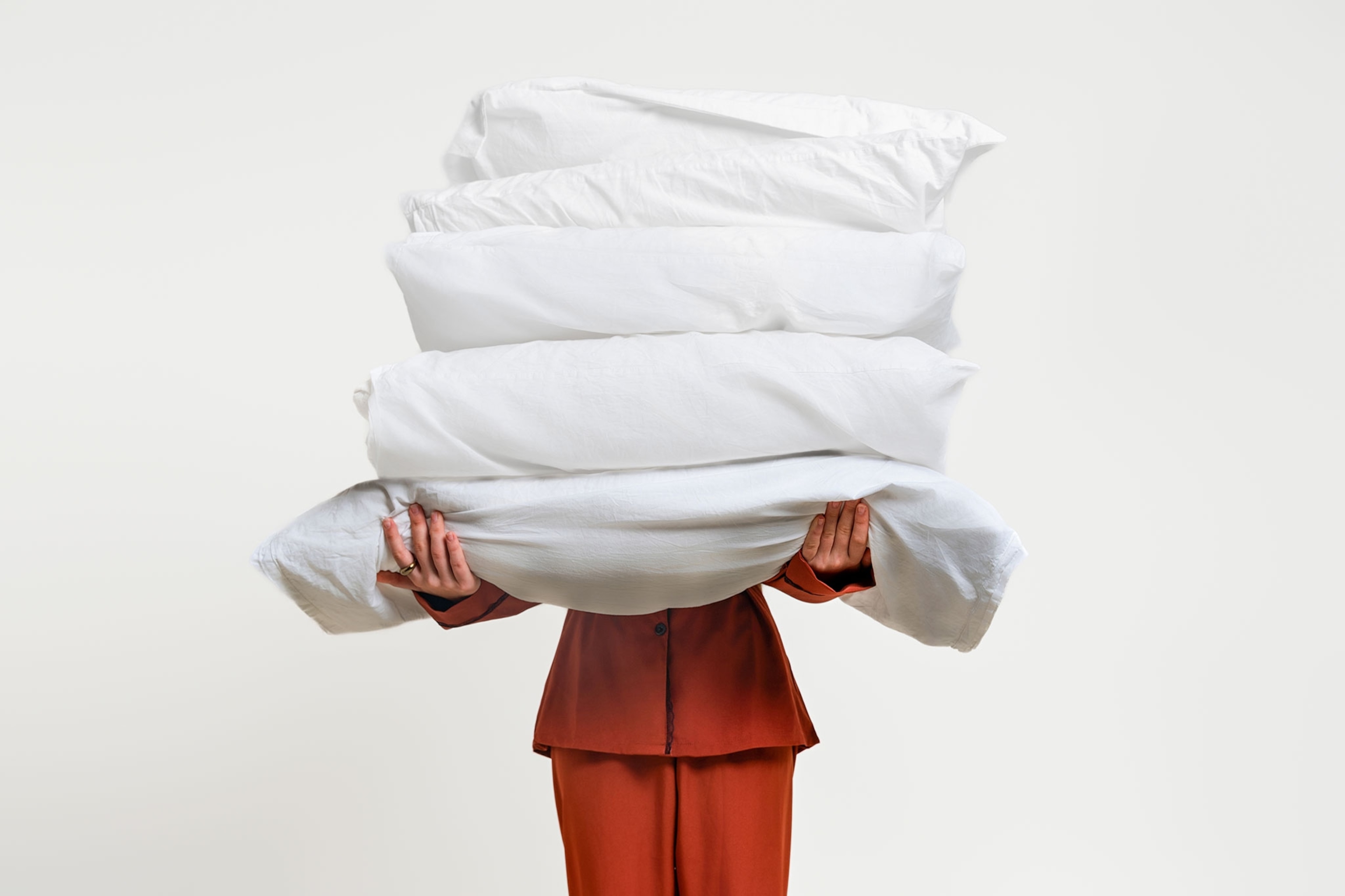 Person in rust-colored pajamas holding a large stack of white pillows, partially obscuring their face, against a white minimalist background.