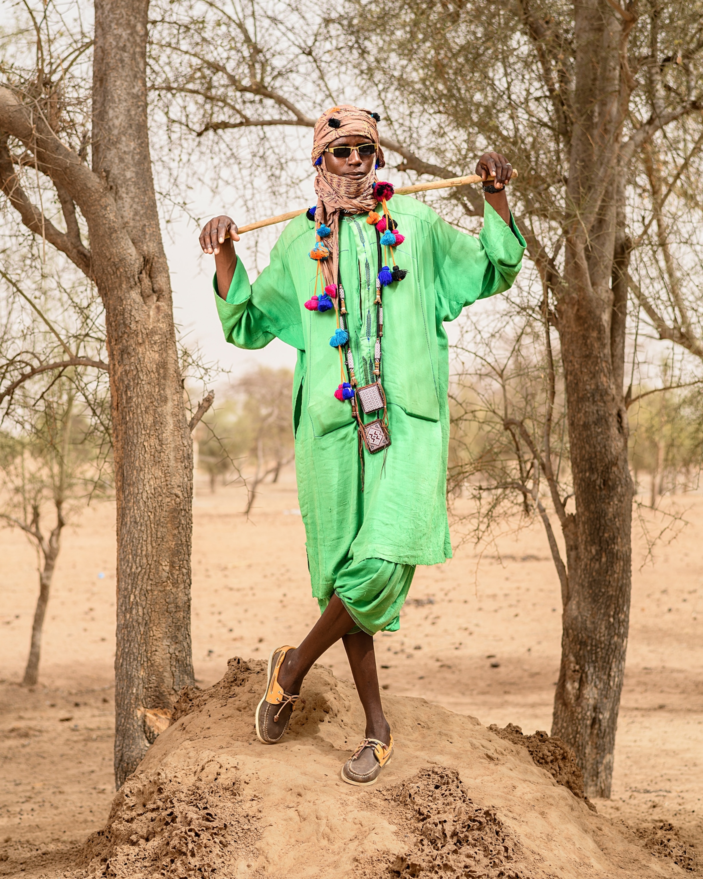 A man in bright green attire and a head wrap stands on a mound of dirt between two trees while holding a stick on his shoulders with both hands.