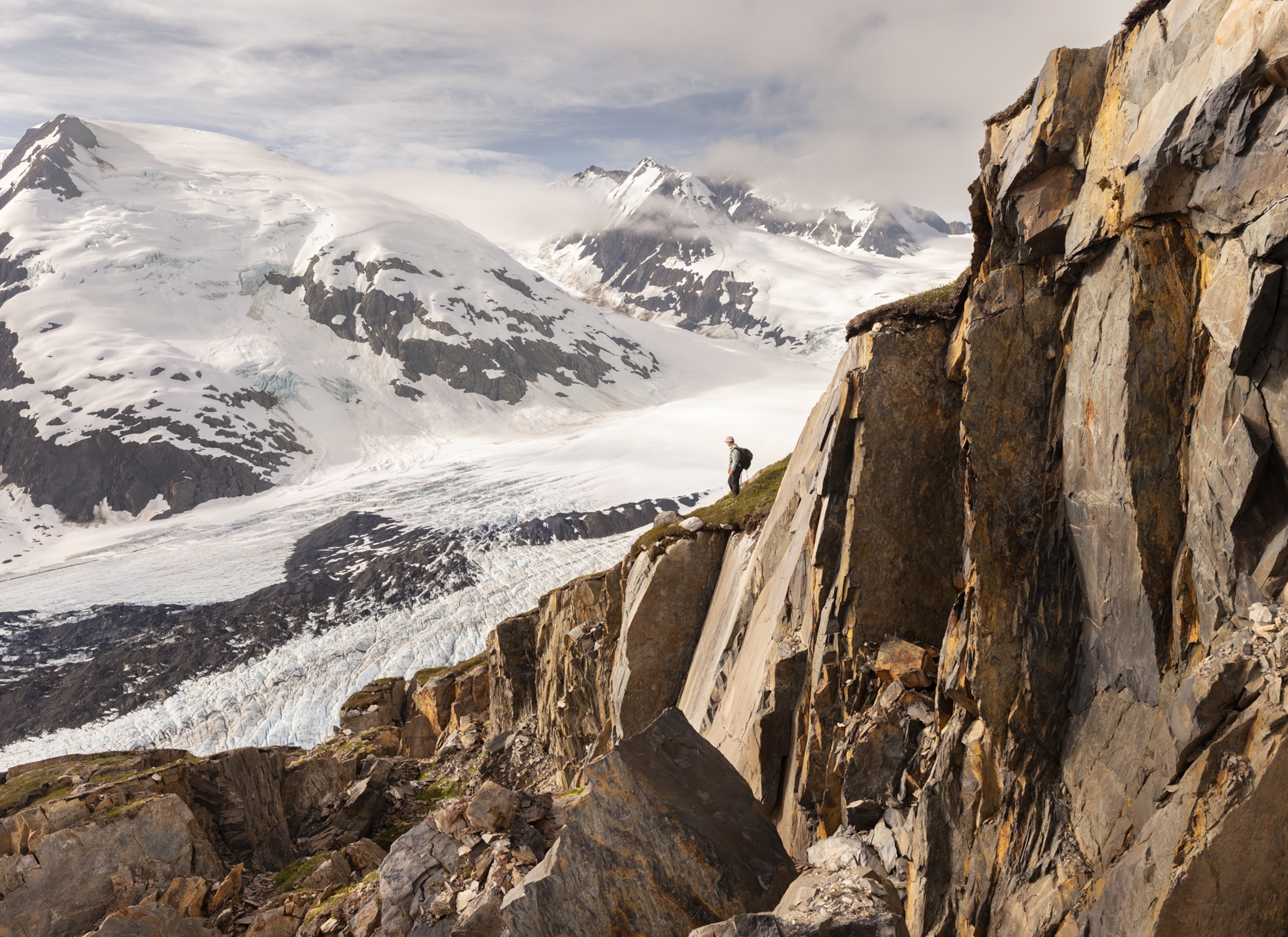 A person stands on a rocky cliff above a wide glacier, with large snowy mountains behind.