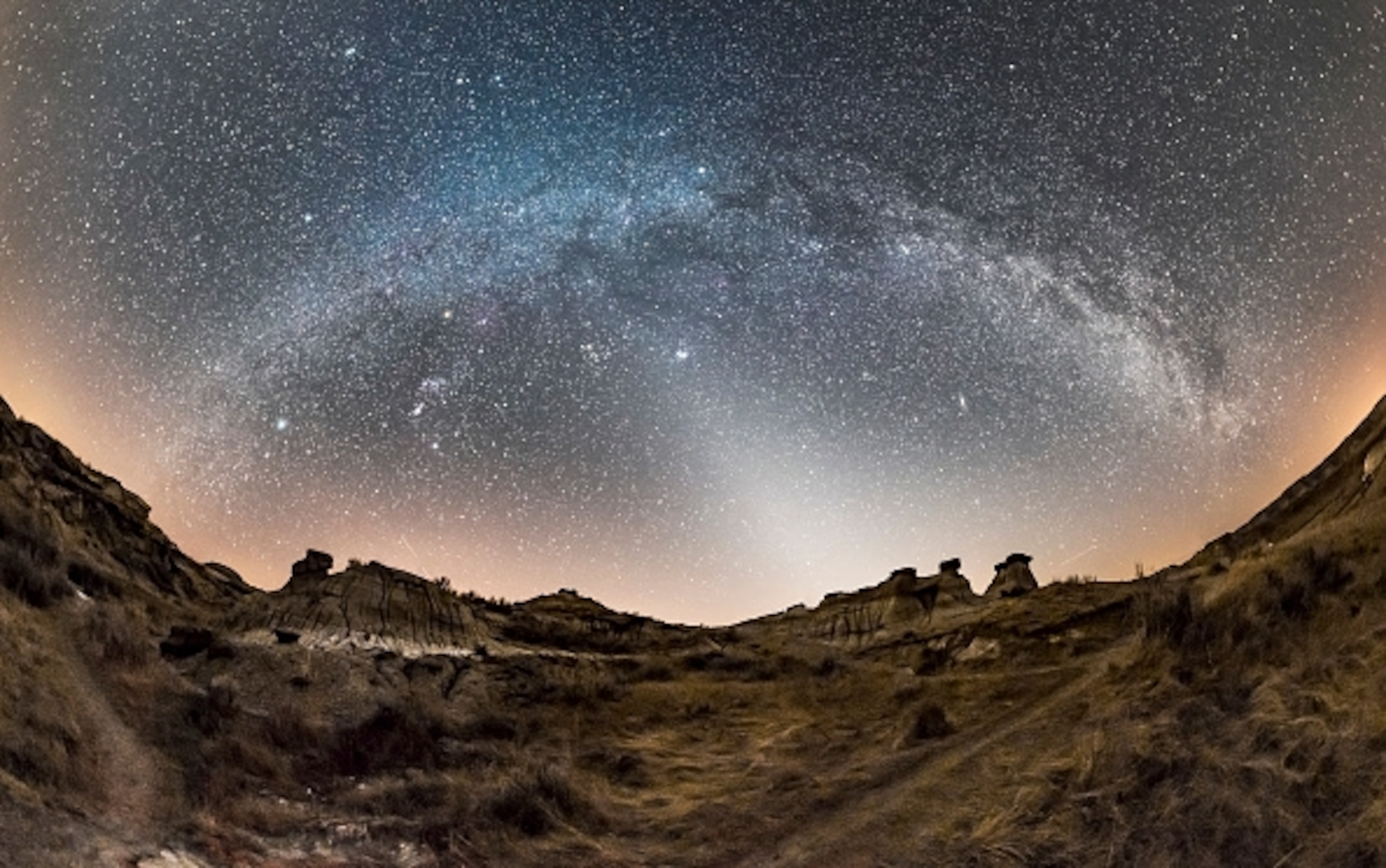 Starry night sky over a desert landscape with rugged hills. The Milky Way forms a bright, arching band across the sky.