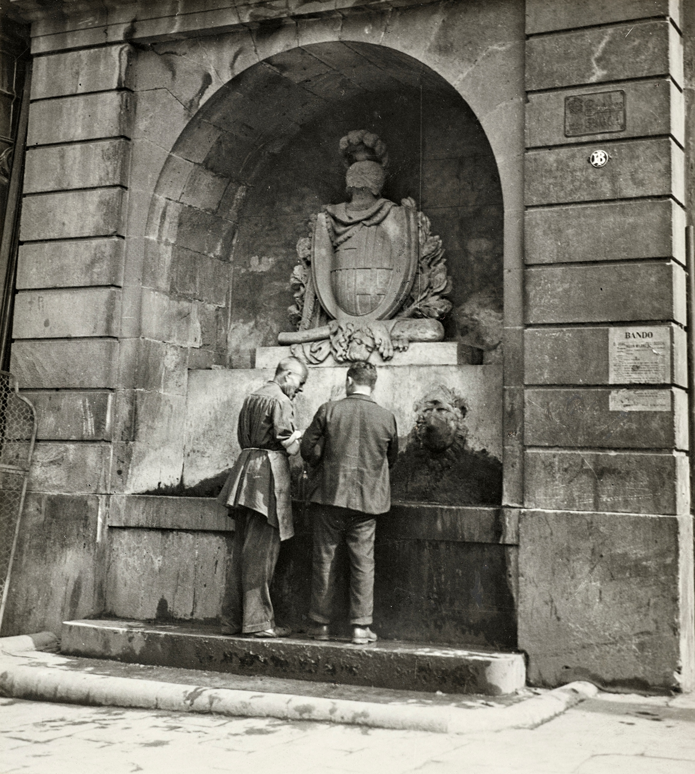 A rear view two men in front of the fountain, Font del Pla de la Boqueria, made in the 19th century, on Place Boqueria (Boqueria Square) at the corner of La Rambla.