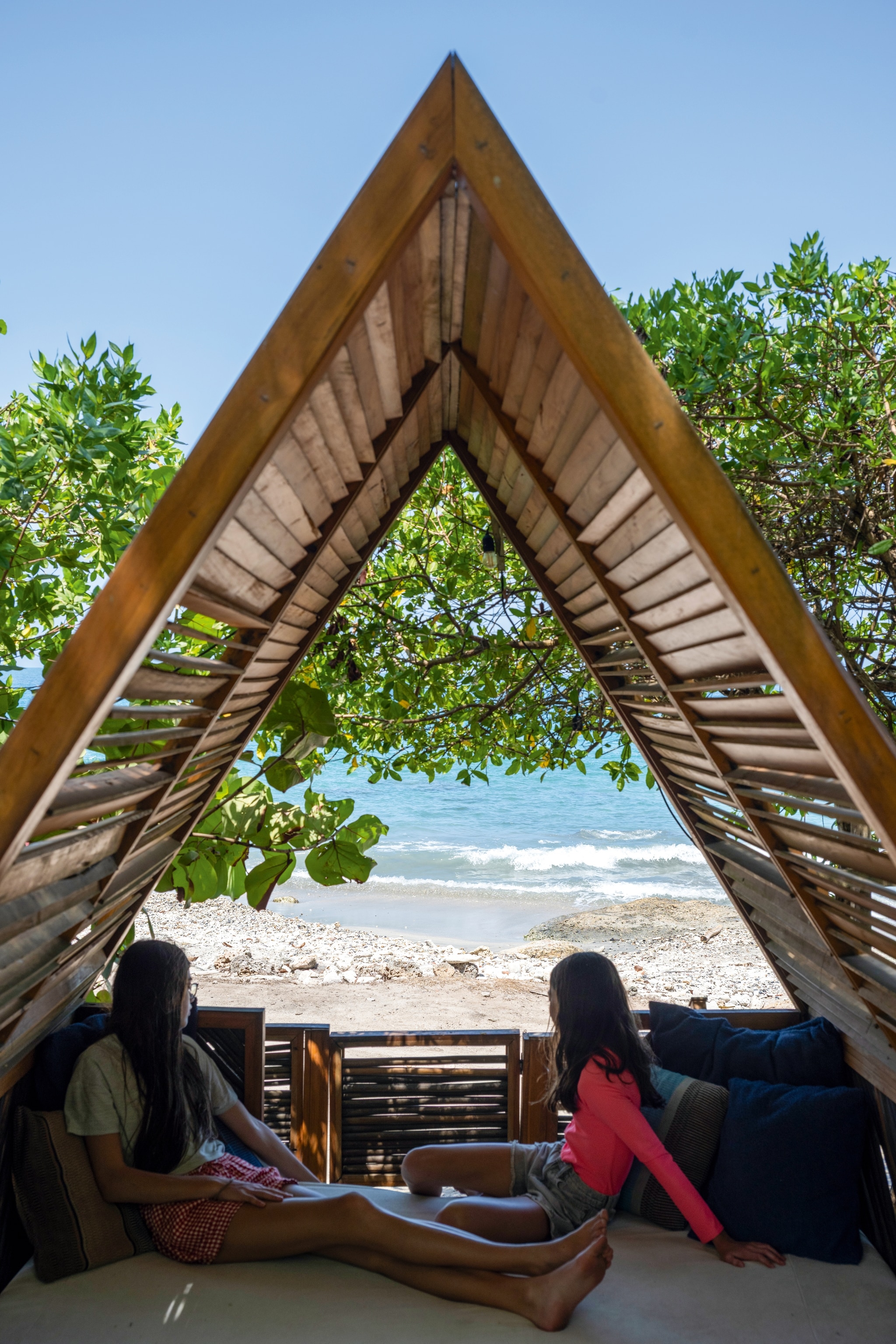 Two people sit under wooden tent looking out at beach
