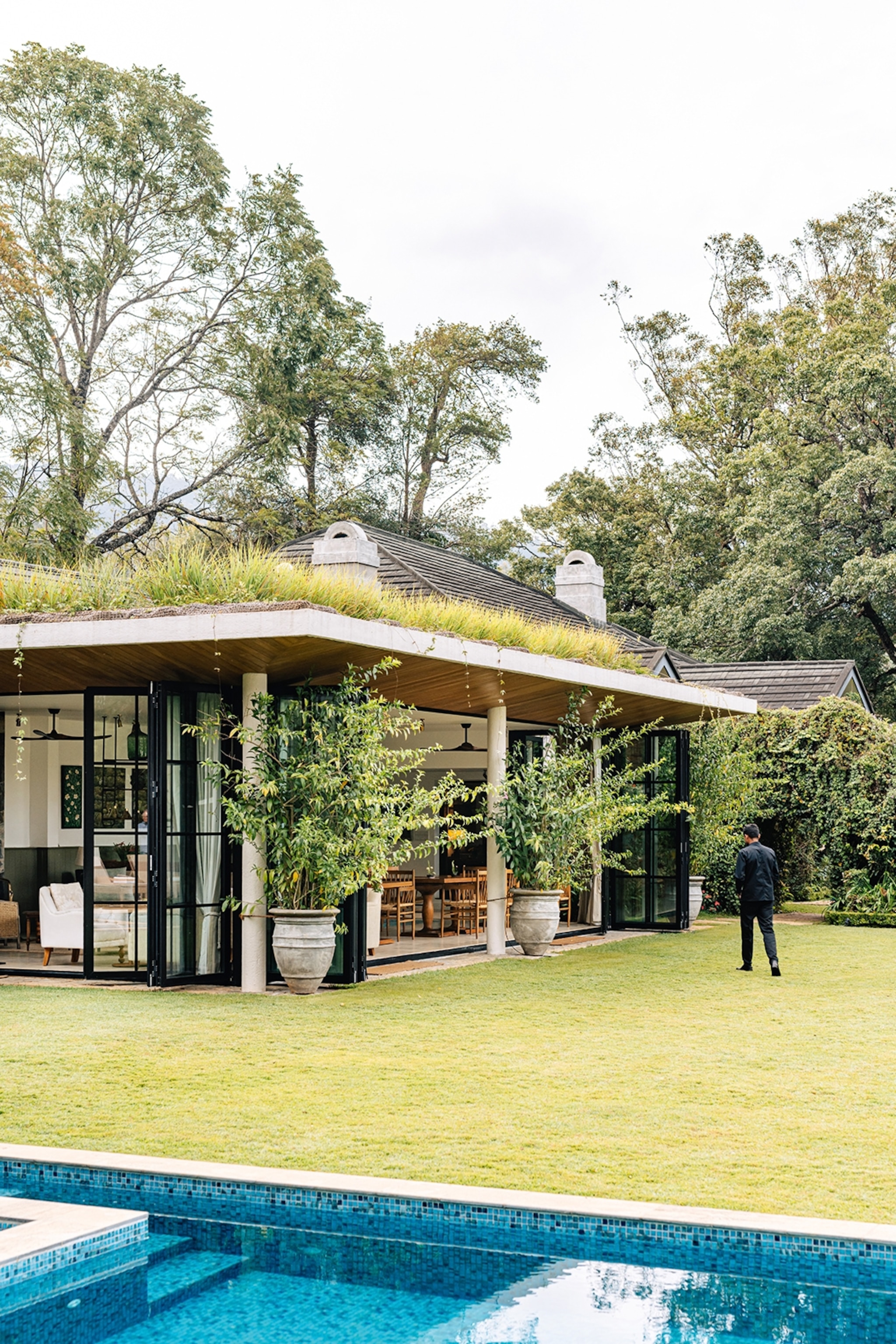 A modernist bungalow with moss growing on one side of its roof, supported by 80s-style stone pilars and featuring floor-to-ceiling windows.