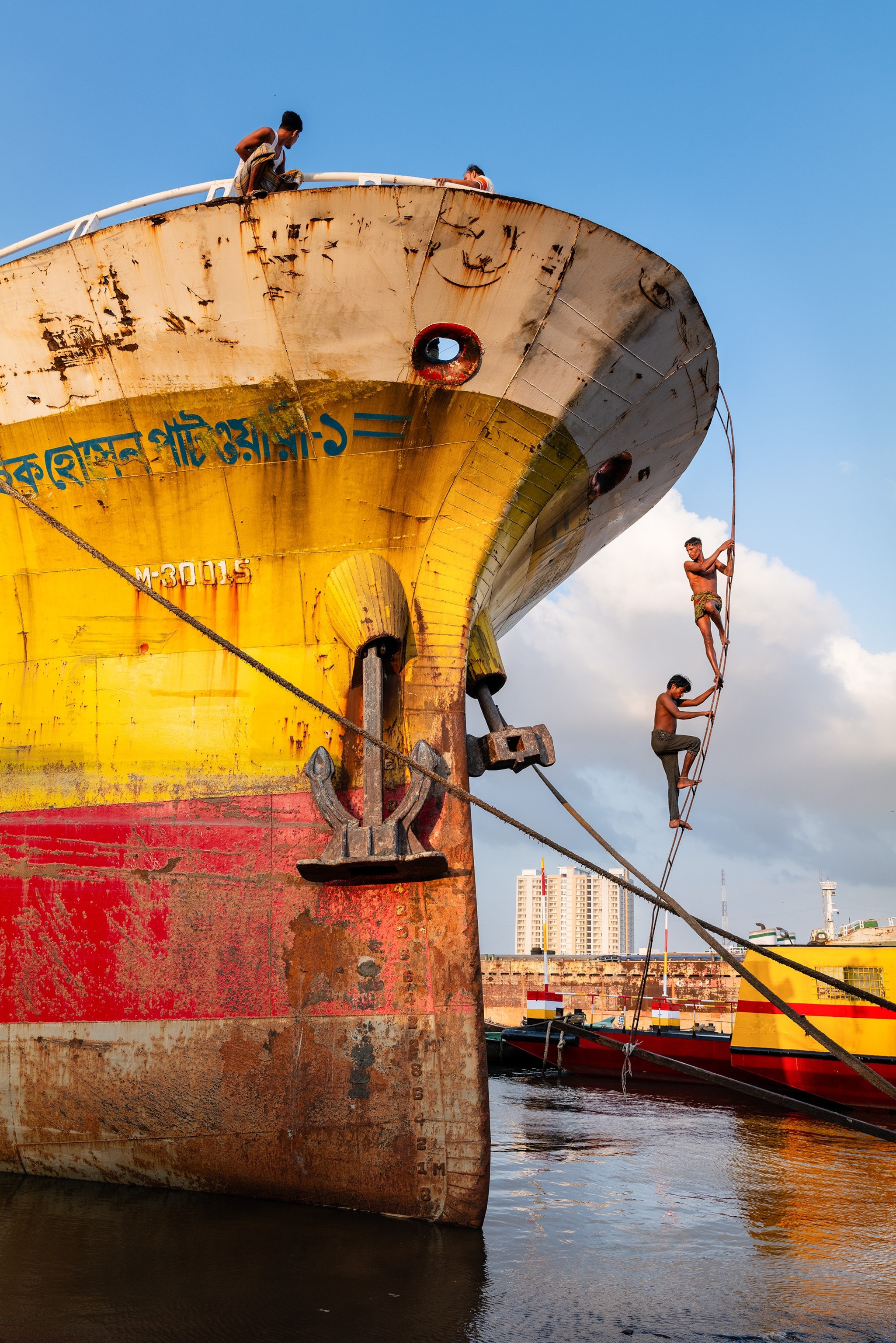 Two shirtless men climb a ladder up to the top of a rusty white, yellow, and red ship.