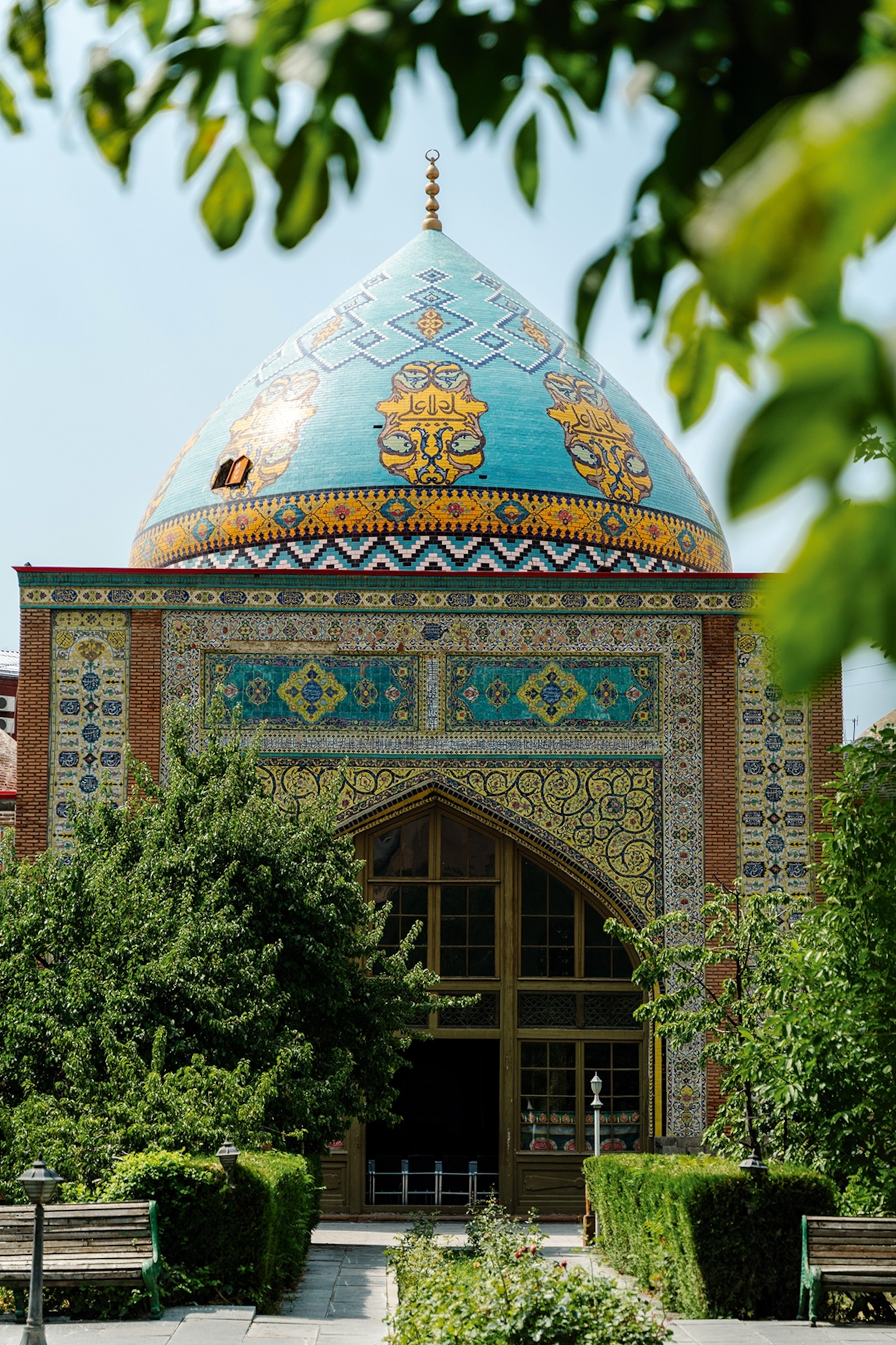 A tiled and domed mosque peeking through the green leaves of a lush garden.
