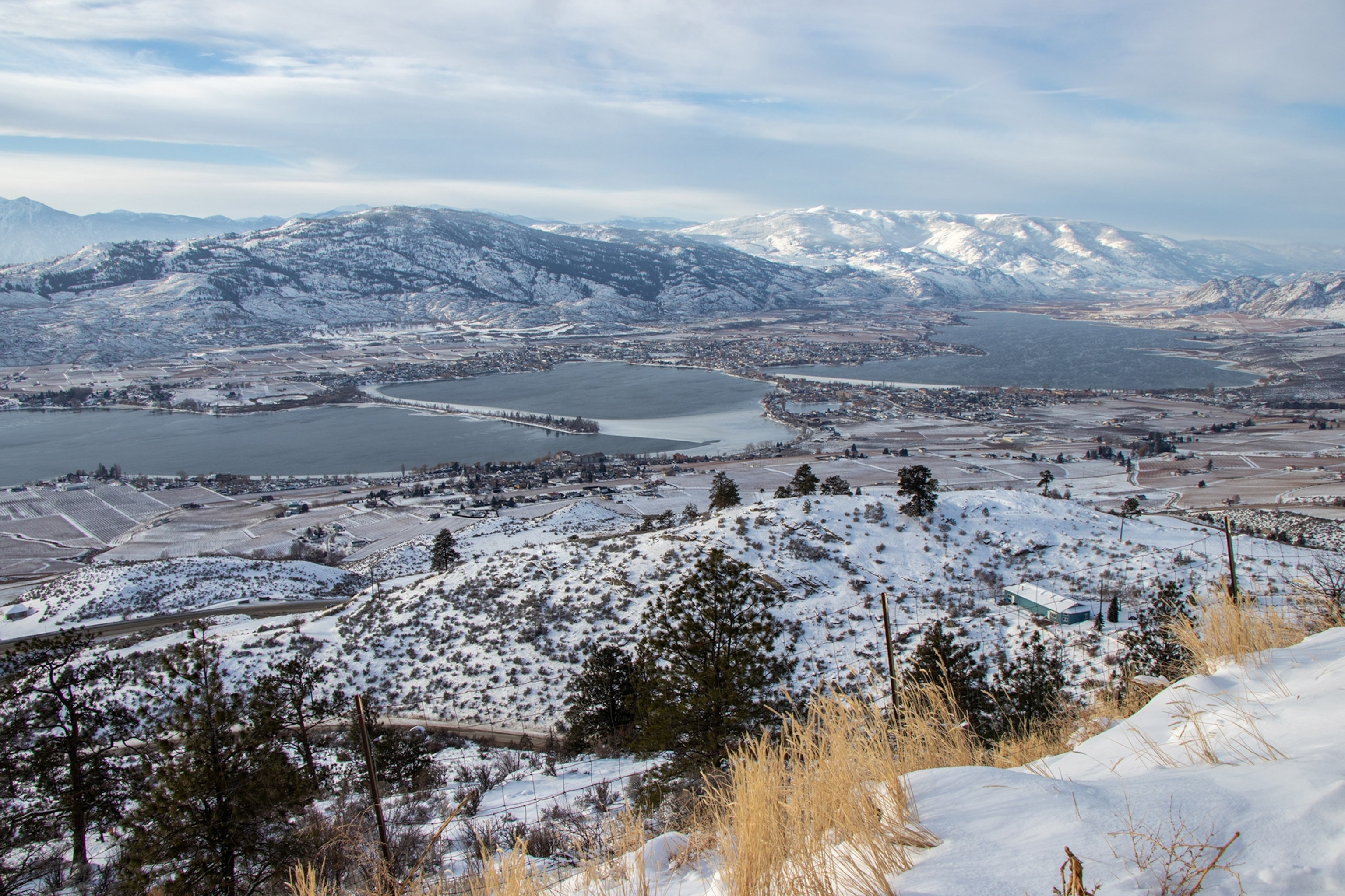 Wide shot from high vantage point looking down onto a snowy desert landscape and lake below, mountains in the distance, cloudy sky.