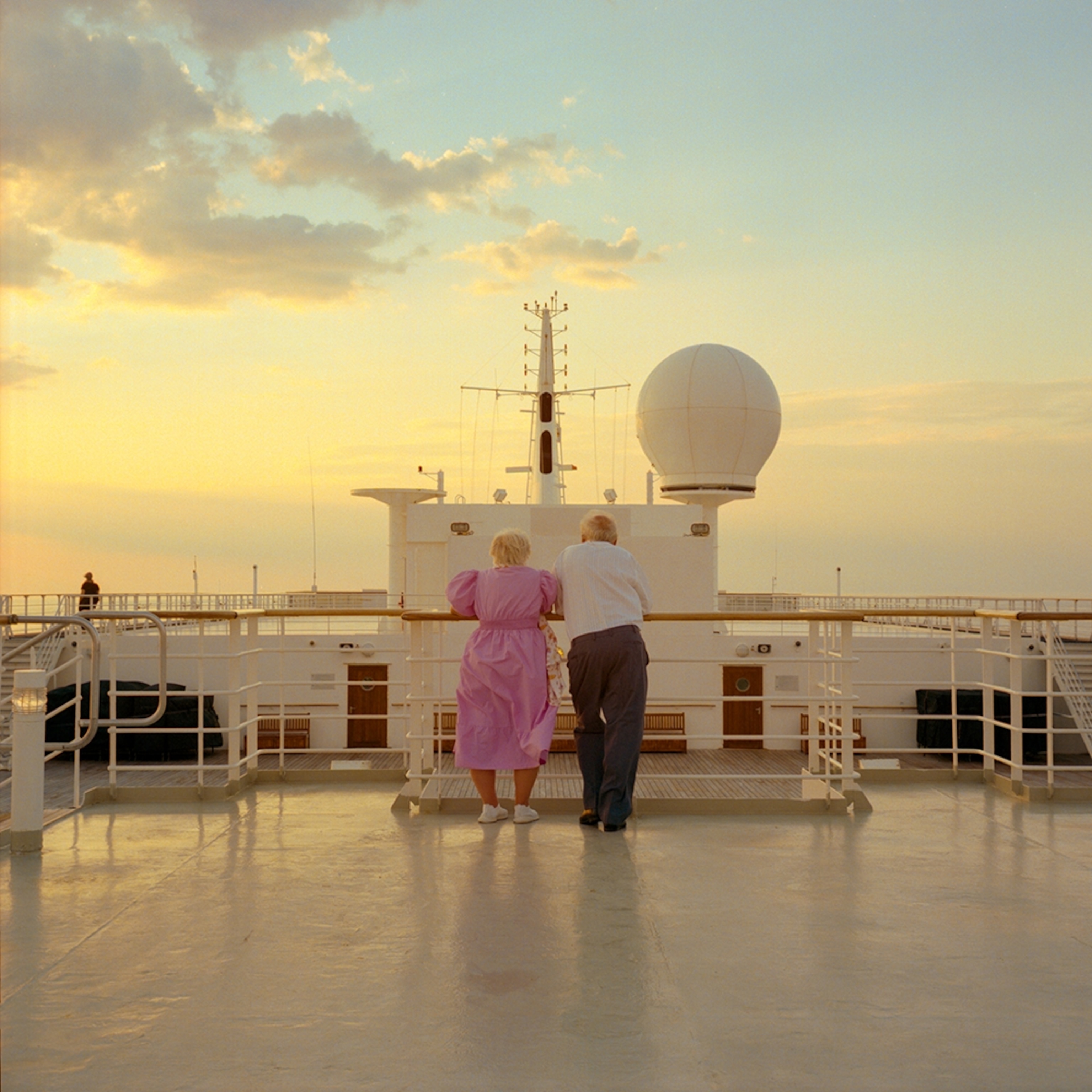 An elderly couple standing on the railing of a cruise ship at sunset.