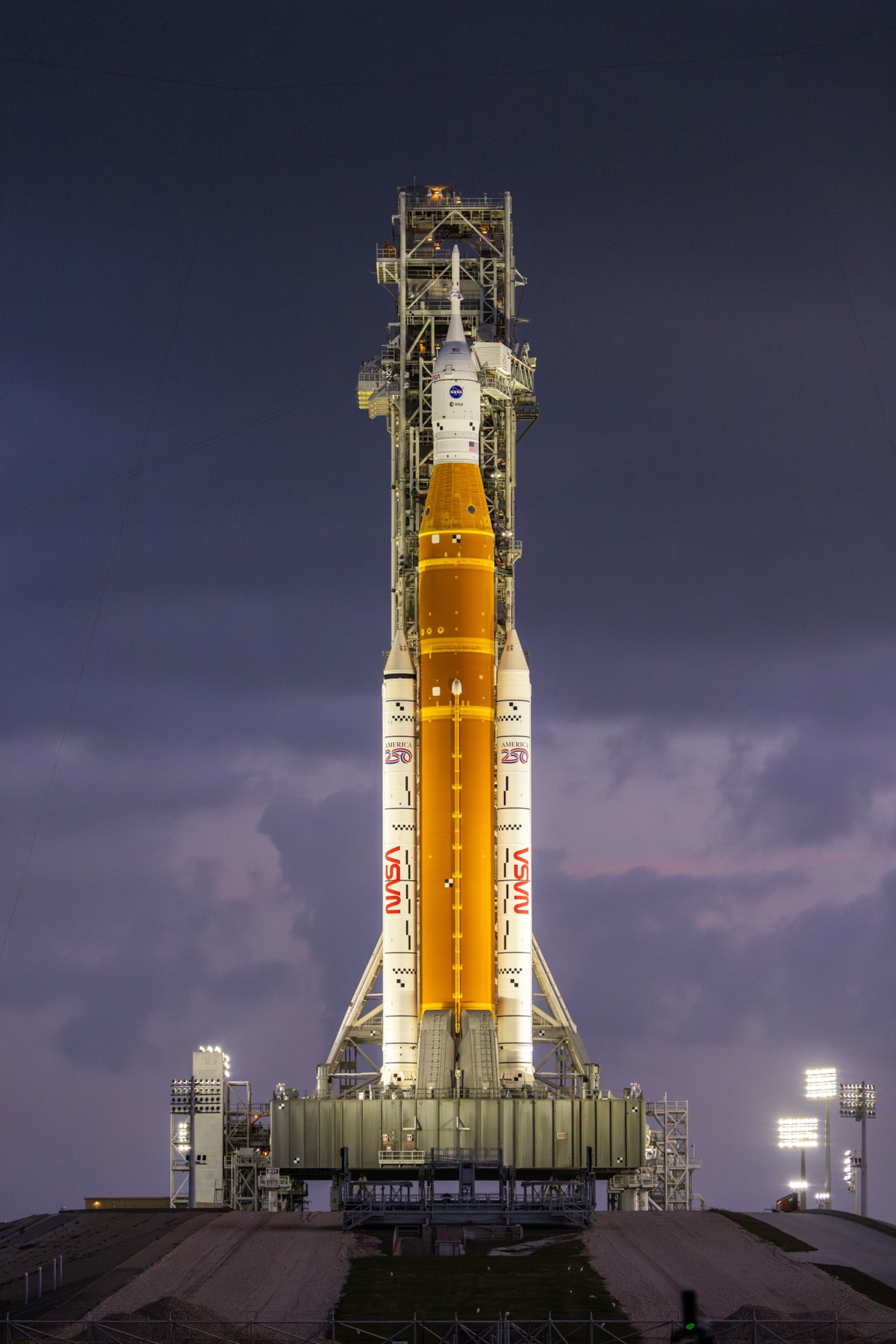 A large rocket with an orange central core and two white boosters stands on a launch pad at dawn, under a cloudy sky. Bright floodlights illuminate the scene