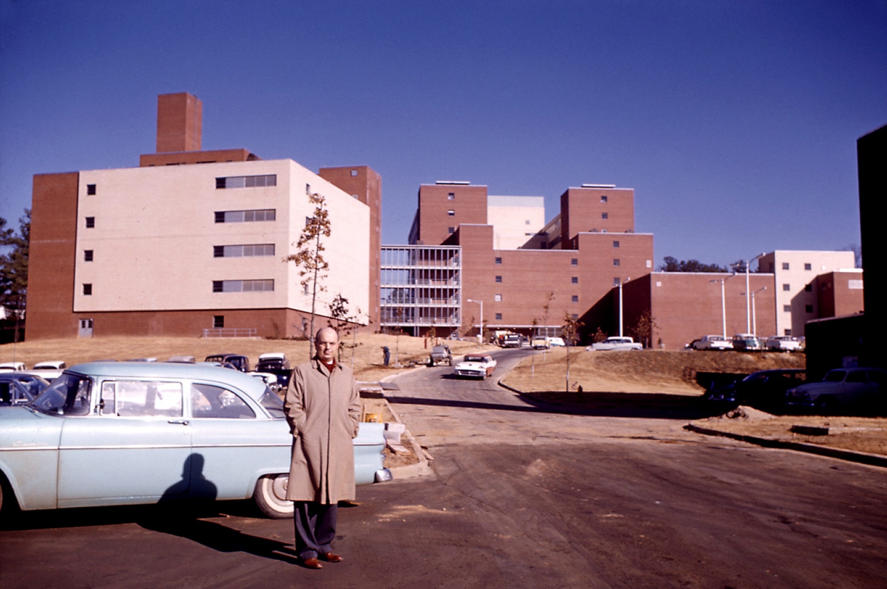 A bald man in a trenchcoat standing for a portrait outside a newly constructed brick building in 1961.