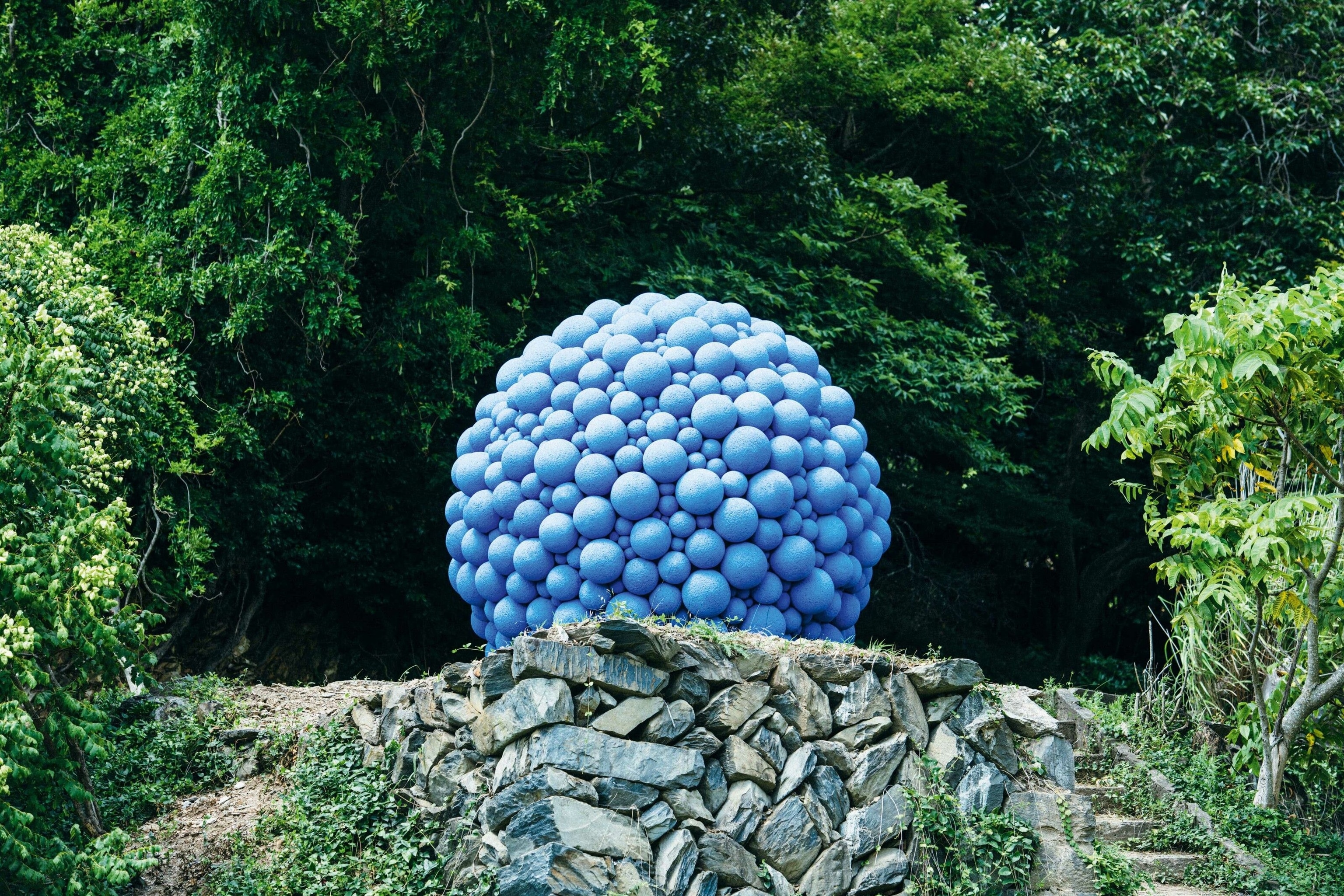Naoki Kusumi's Bubble sits atop a mound of stones, surrounded by woodland. It is a unique piece with a spherical shape, comprised of hundreds of smaller blue balls.
