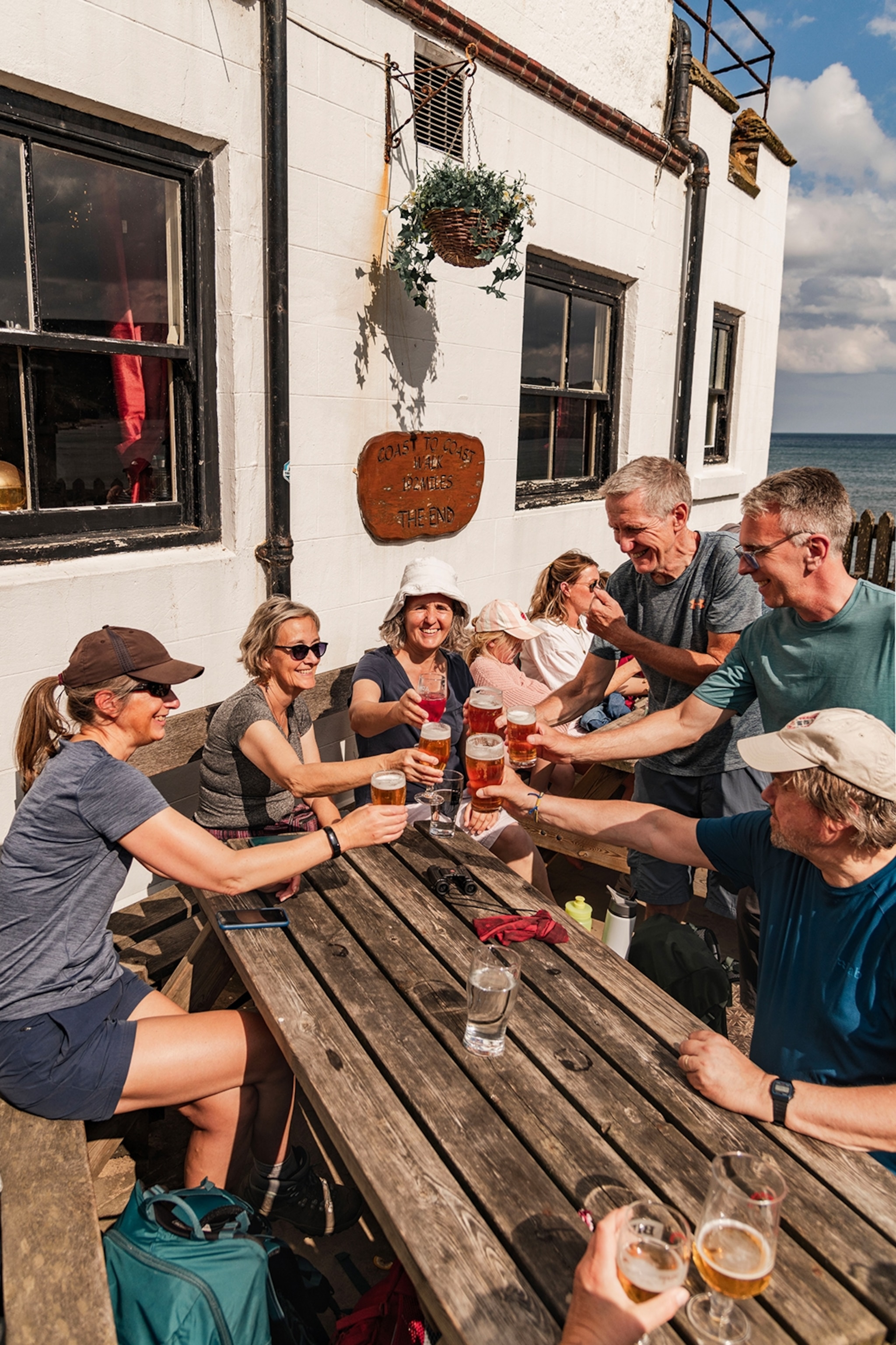 A group of middle-aged hikers cheering with pints of beer on a pub terrace in the sun.