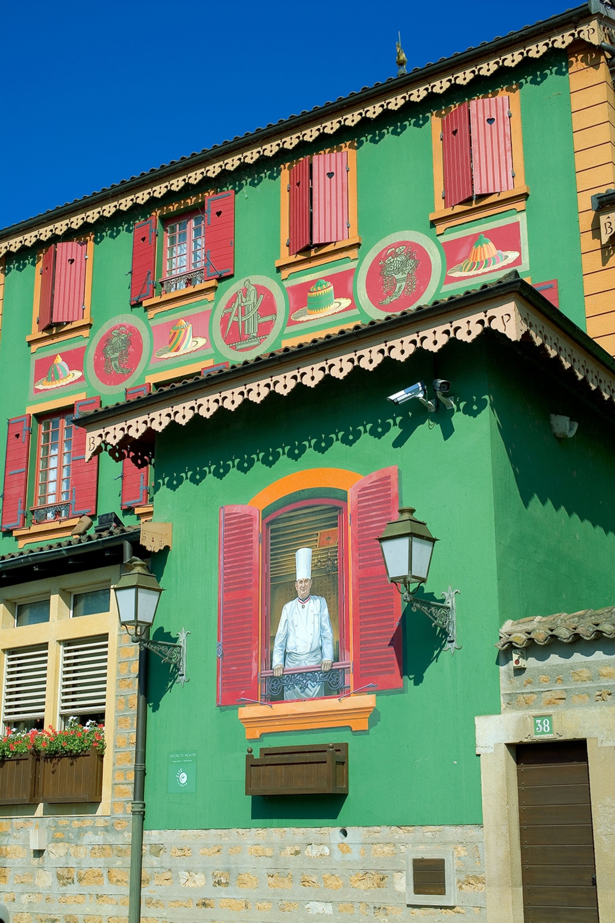 The painted fassade of a restaurant depicting a classic chef looking out through windows with wooden shutters.