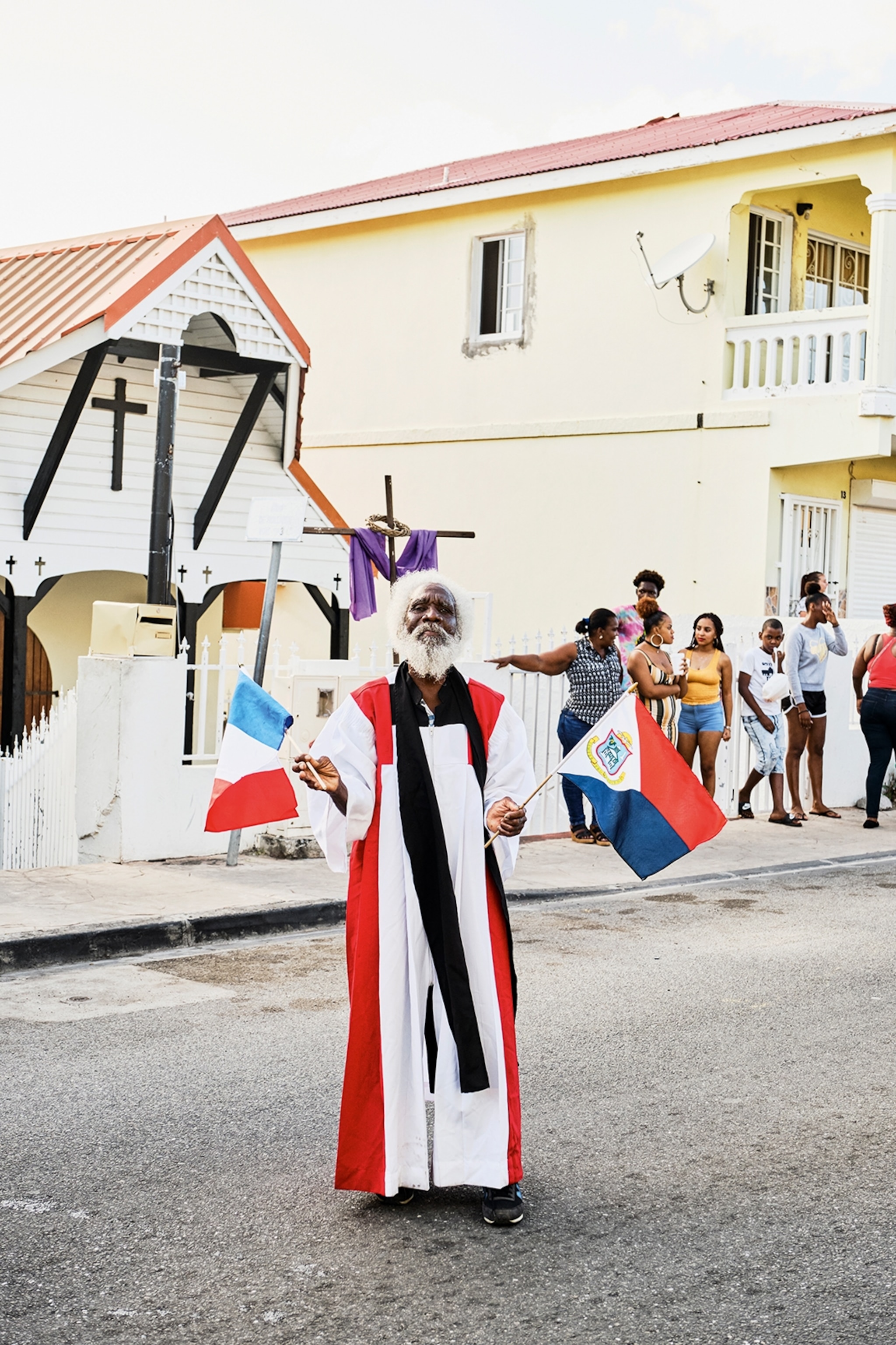 An elderly man standing in the middle of a street wearing a long priest dress and waving Caribbean flags.
