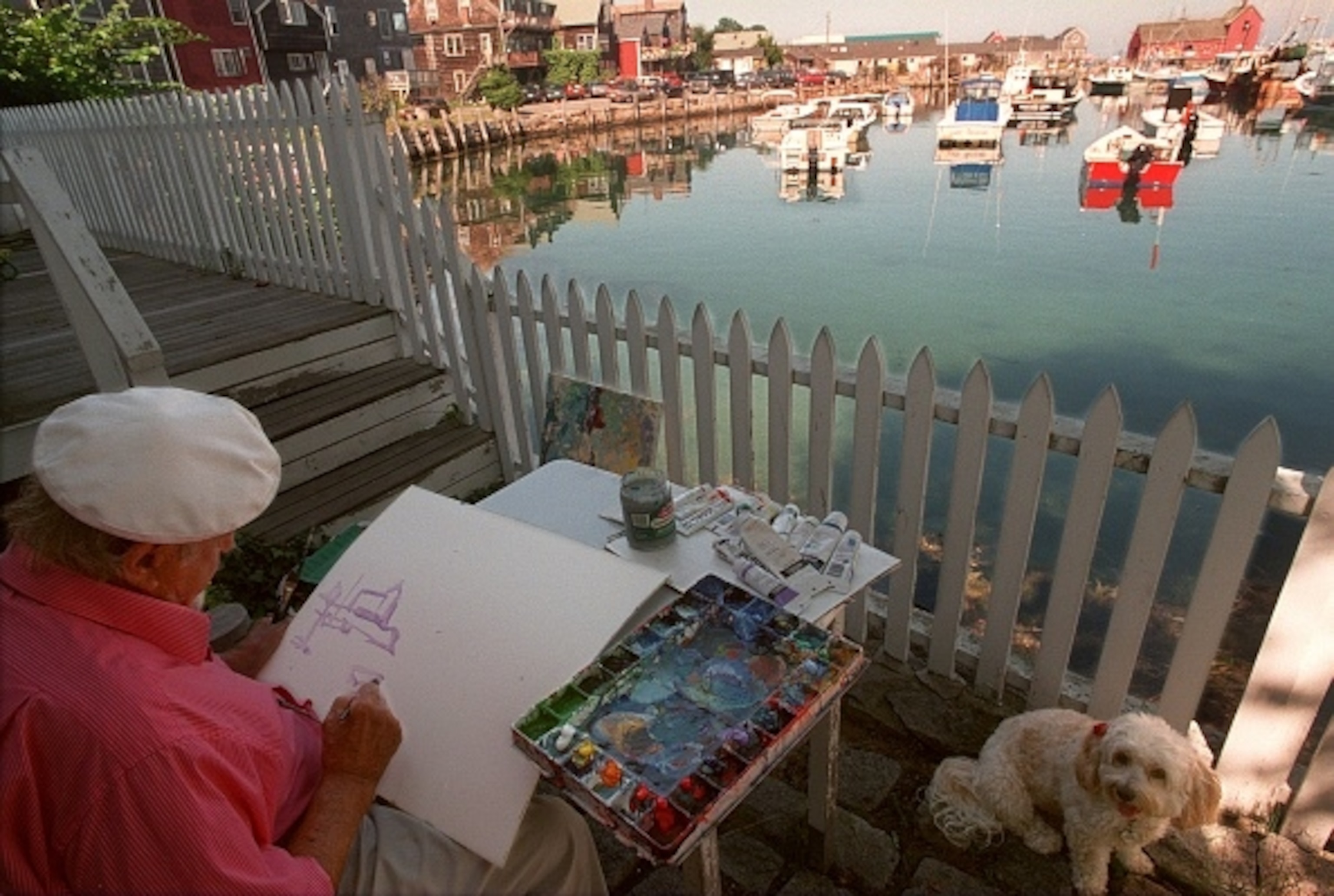 A man sits on a dock on the ocean, as boats pass by while painting the scene in front of him