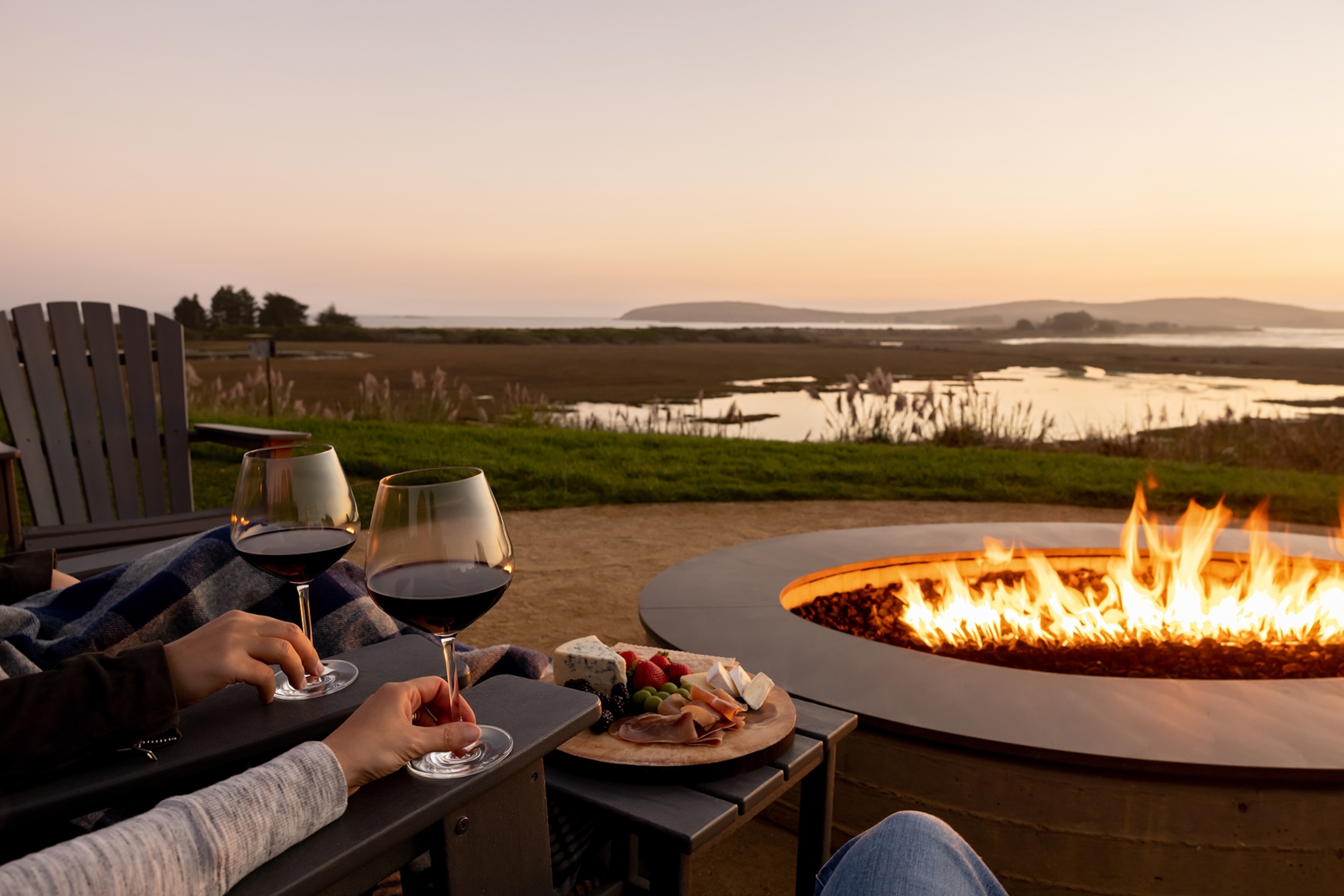 A sunset scene of a couple holding up two glasses of red wine next to a fire pit, looking out at the lake-dotted landscape ahead.